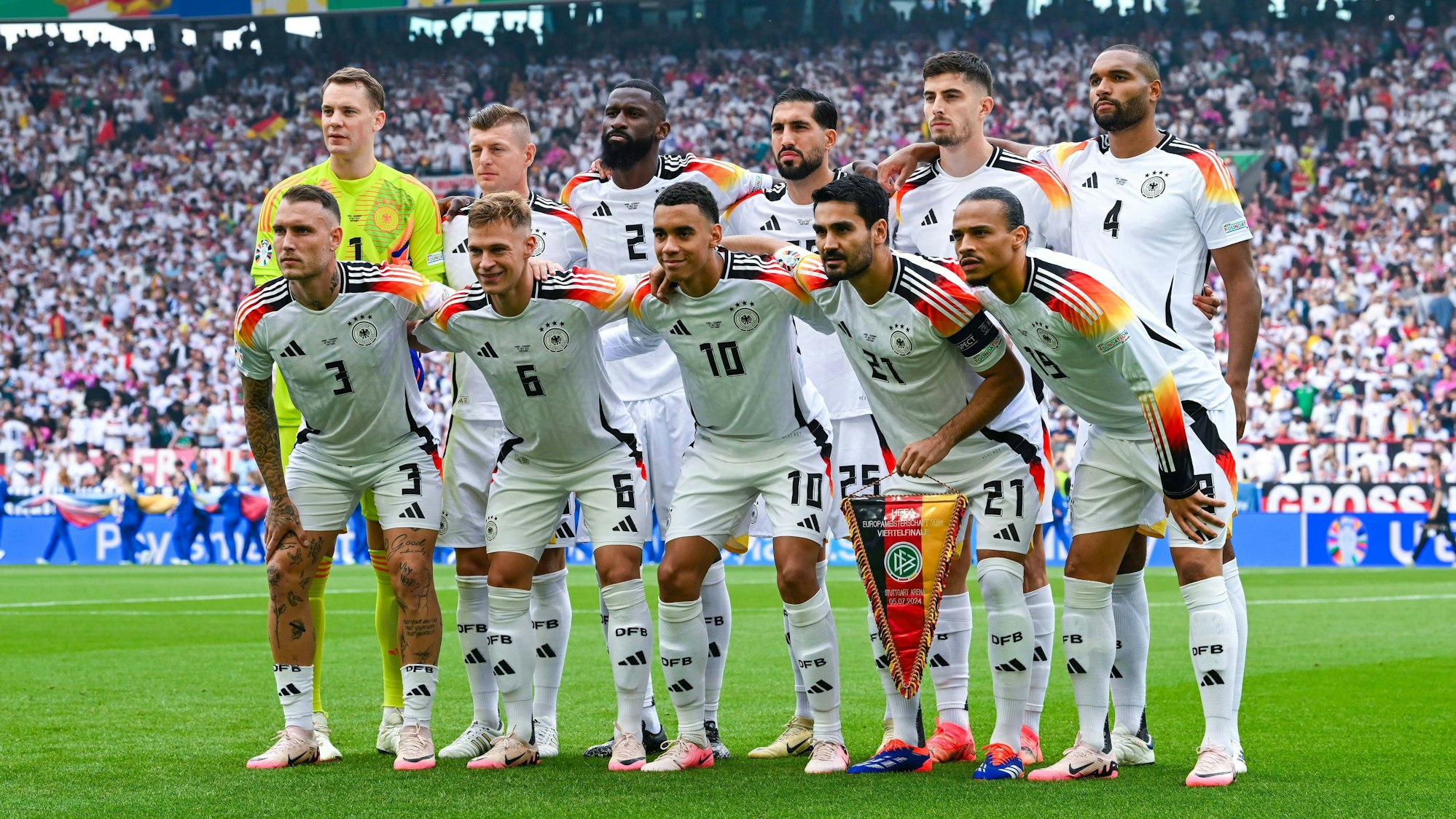 Die deutsche Nationalmannschaft posiert in zwei Reihen auf dem Spielfeld: Manuel Neuer, Toni Kroos, Antonio Rüdiger, Emre Can, Kai Havertz, Jonathan Tah, David Raum, Joshua Kimmich, Jamal Musiala, Ilkay Gündogan und Leroy Sané. Im Hintergrund sind voll besetzte Stadionränge zu sehen.