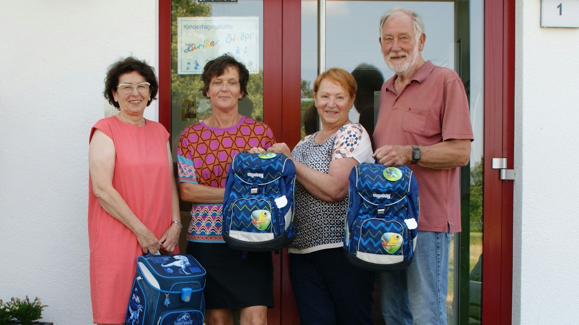 Drei Frauen und ein Mann stehen mit Schulranzen in den Händen vor der Eingangstür einer Kita.