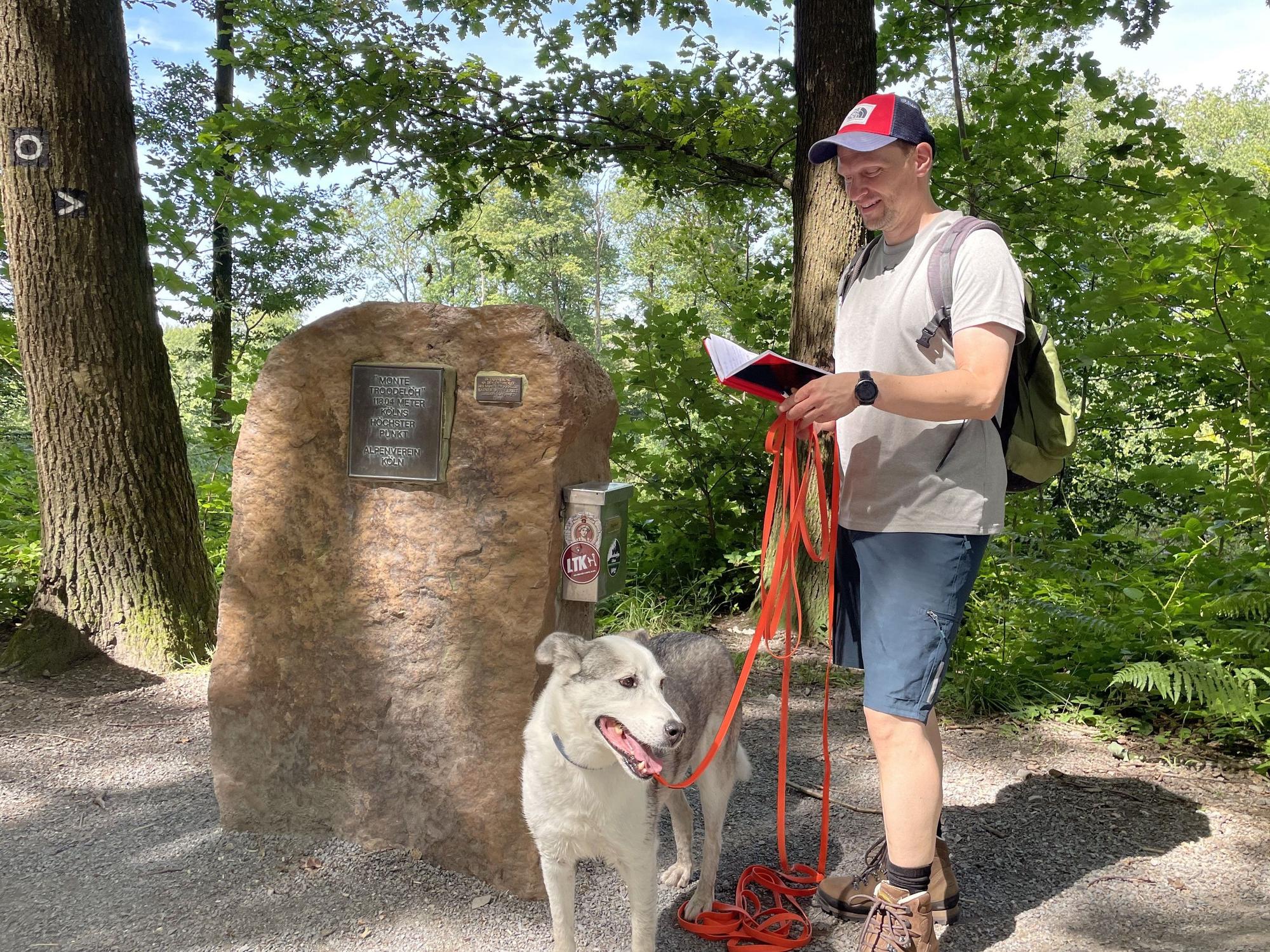 Ein Mann mit Hund steht vor dem Gipfelstein am Monte Troodelöh, dem höchsten Berg Kölns.