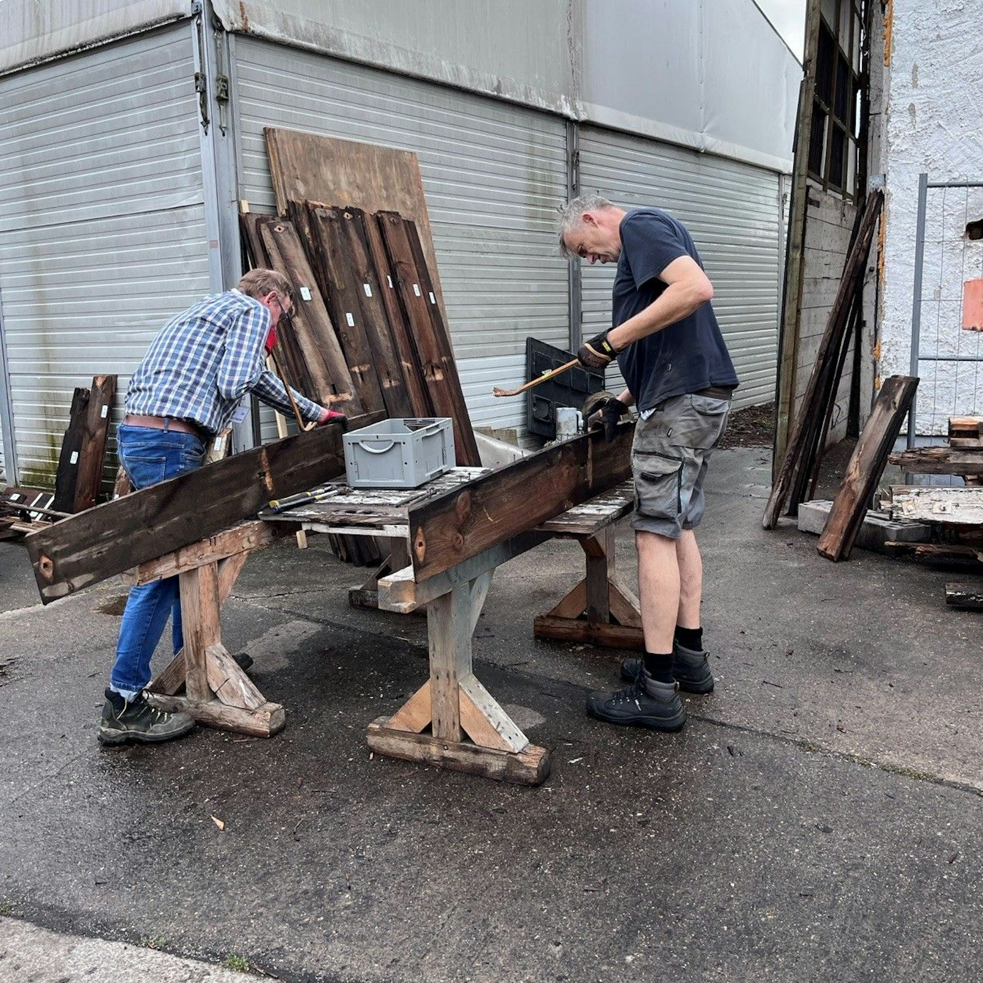 Zwei Männer arbeiten an Holzbalken, die auf Arbeitsböcken liegen.