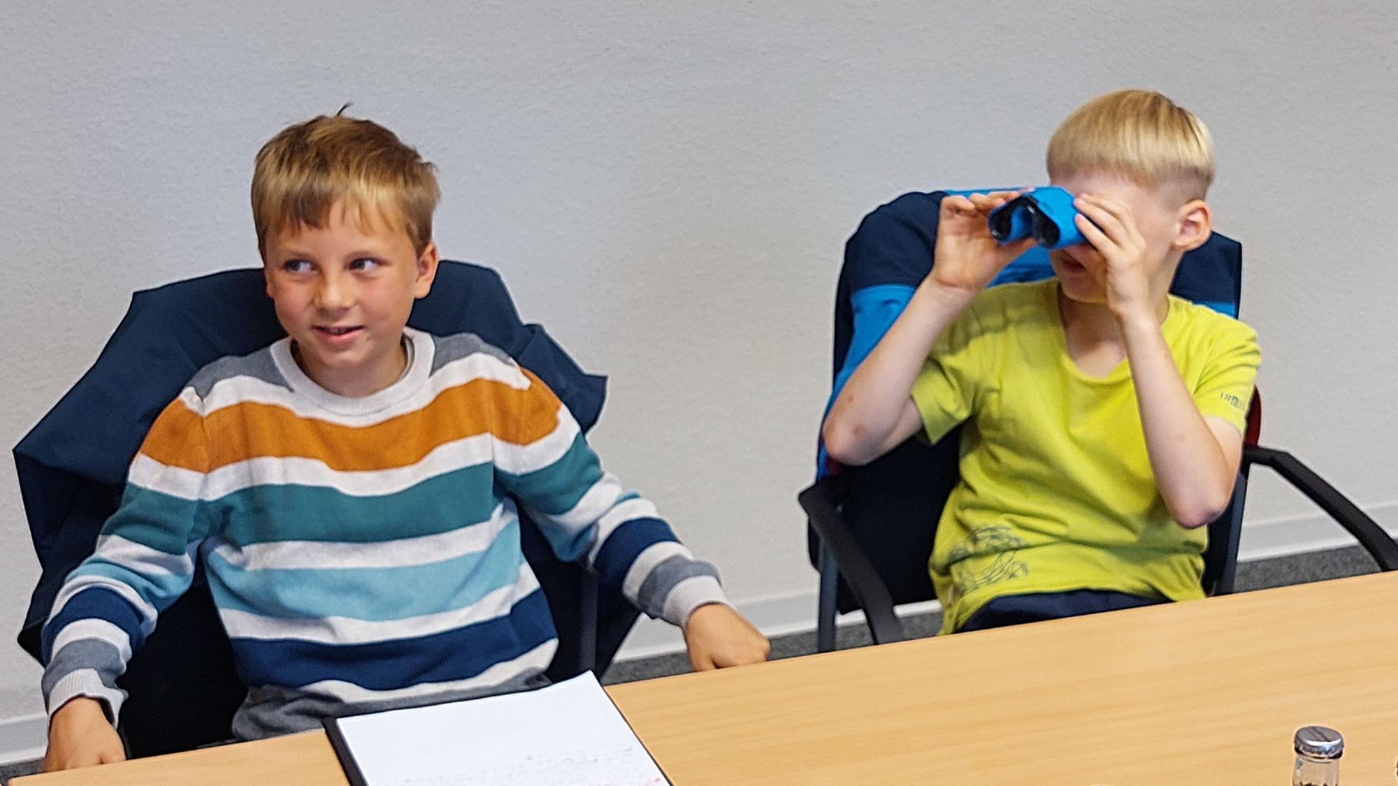 Zwei Jungen sitzen auf Stühlen an einem Tisch. Einer guckt durch ein Fernglas.