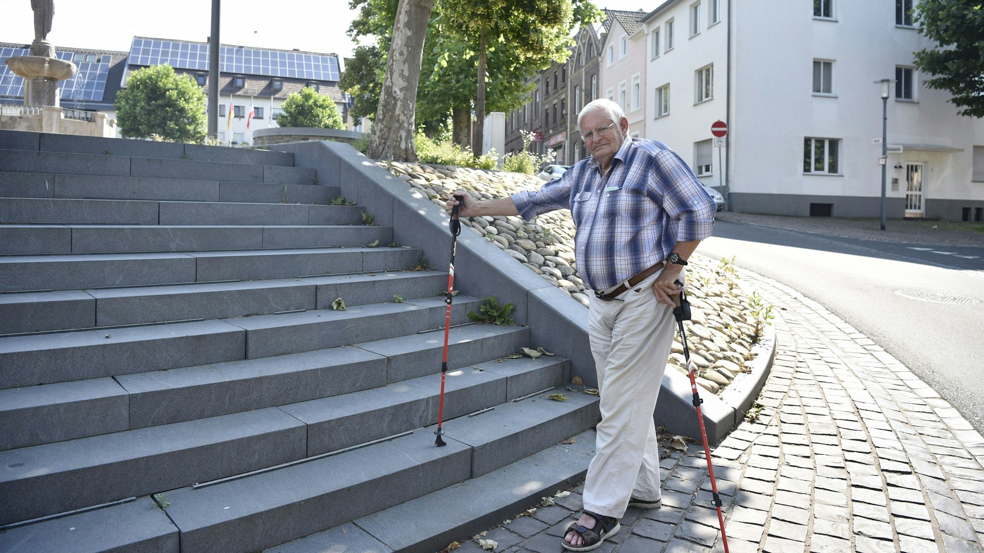 Wolfgang Lüttgen steht an einer Treppe, die kein Geländer hat. Er hat in jeder Hand einen Stock und trägt eine helle, lange Hose, ein kariertes Hemd mit hochgekrempelten Ärmeln sowie Sandalen.