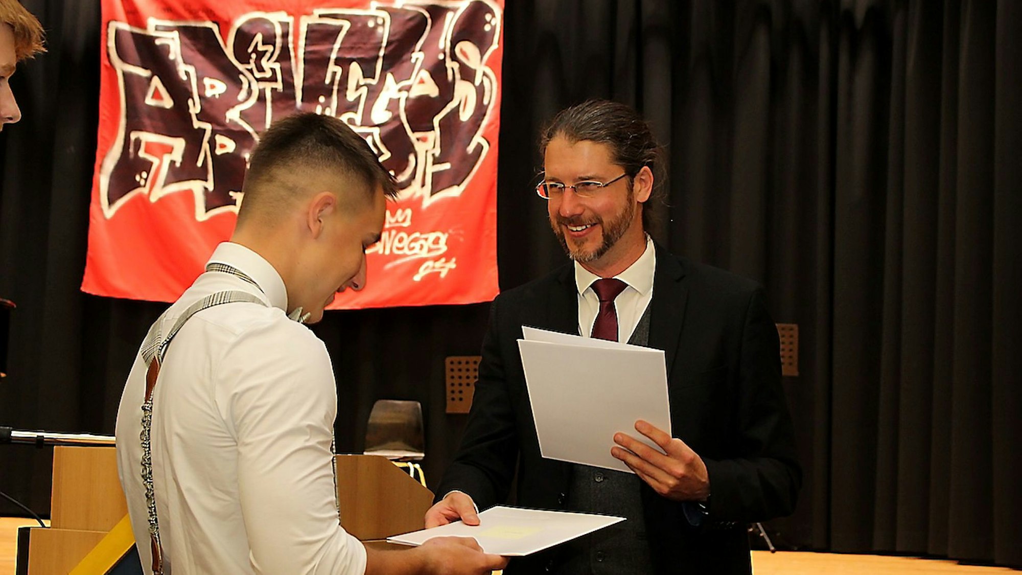 Ein Mann in dunklem Anzug und roter Krawatte überreicht einem jungen Mann in weißem Hemd ein Papier.