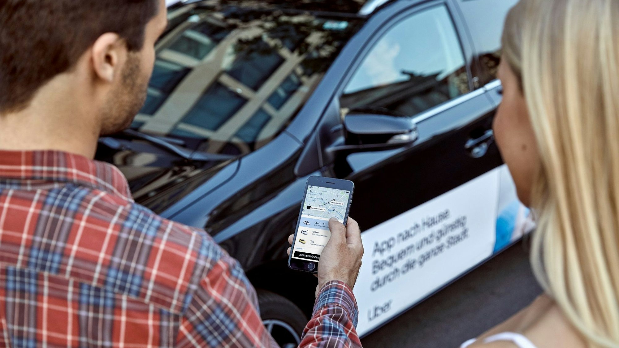 Zwei Personen schauen auf ein Smartphone, auf dem die Uber-App geöffnet ist. Im Hintergrund ein Fahrzeug mit Uber-Beschriftung.