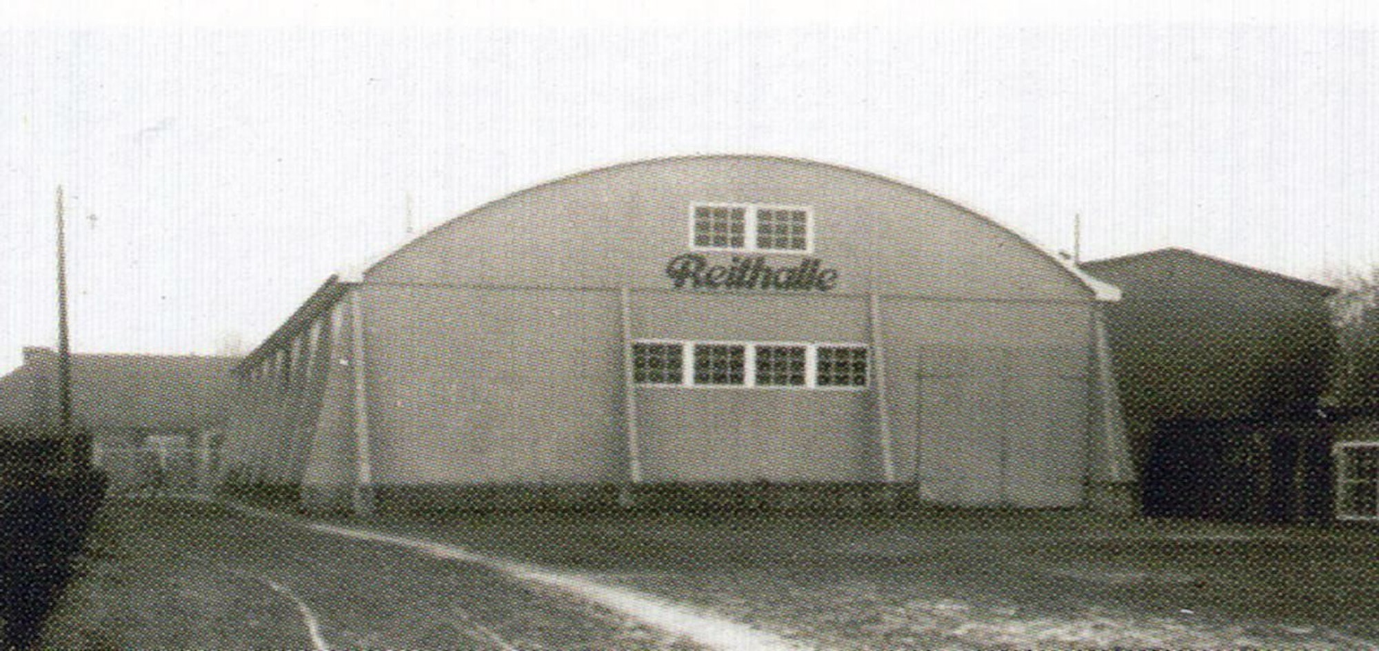 Ein schwarz-weiß Bild zeigt eine Halle mit Walmdach und der Aufschrift Reithalle.