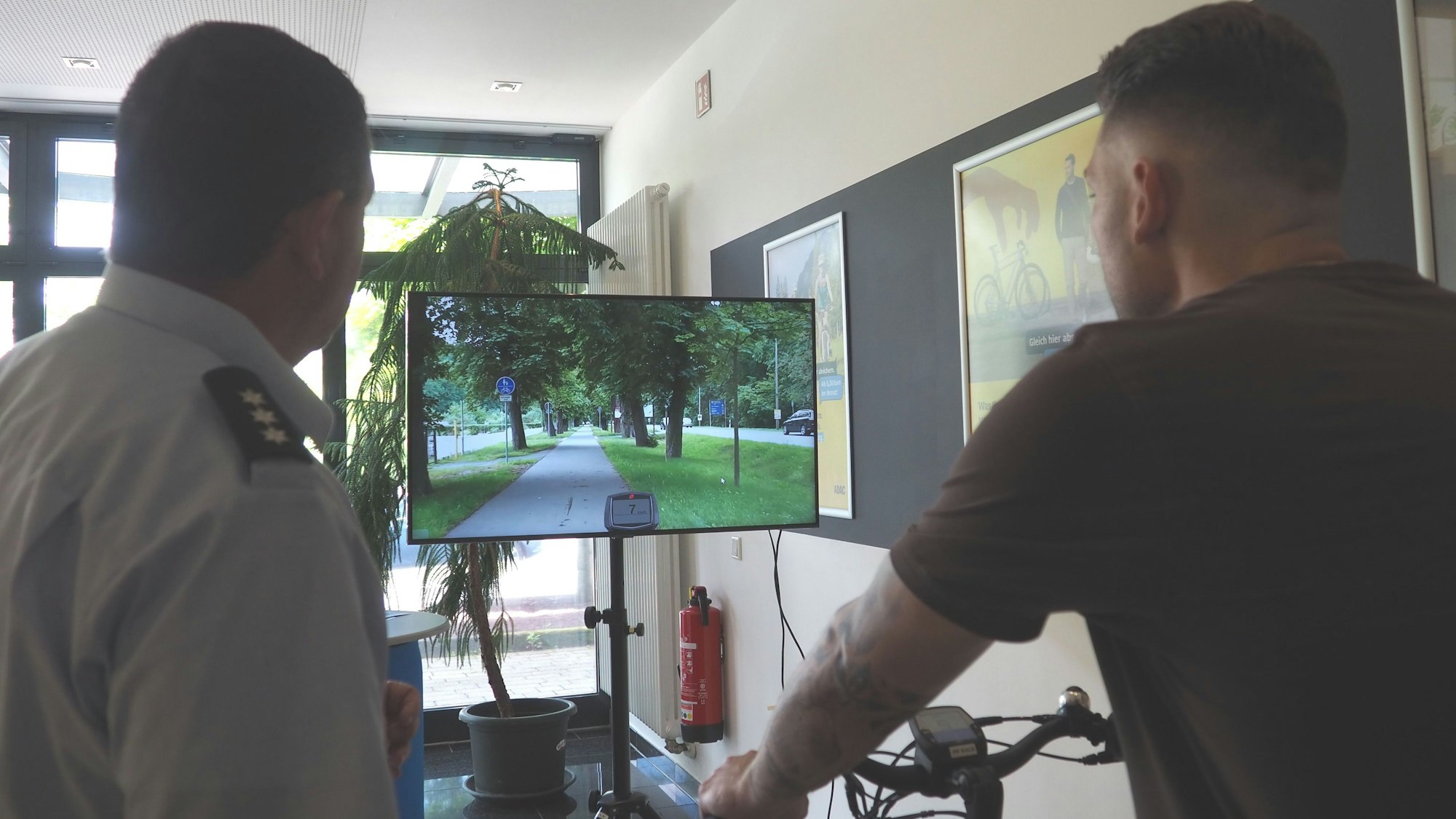 Alexej Loch sitzt auch einem Fahrrad und schaut auf den Bildschirm, neben ihm steht Jörg Meyer.