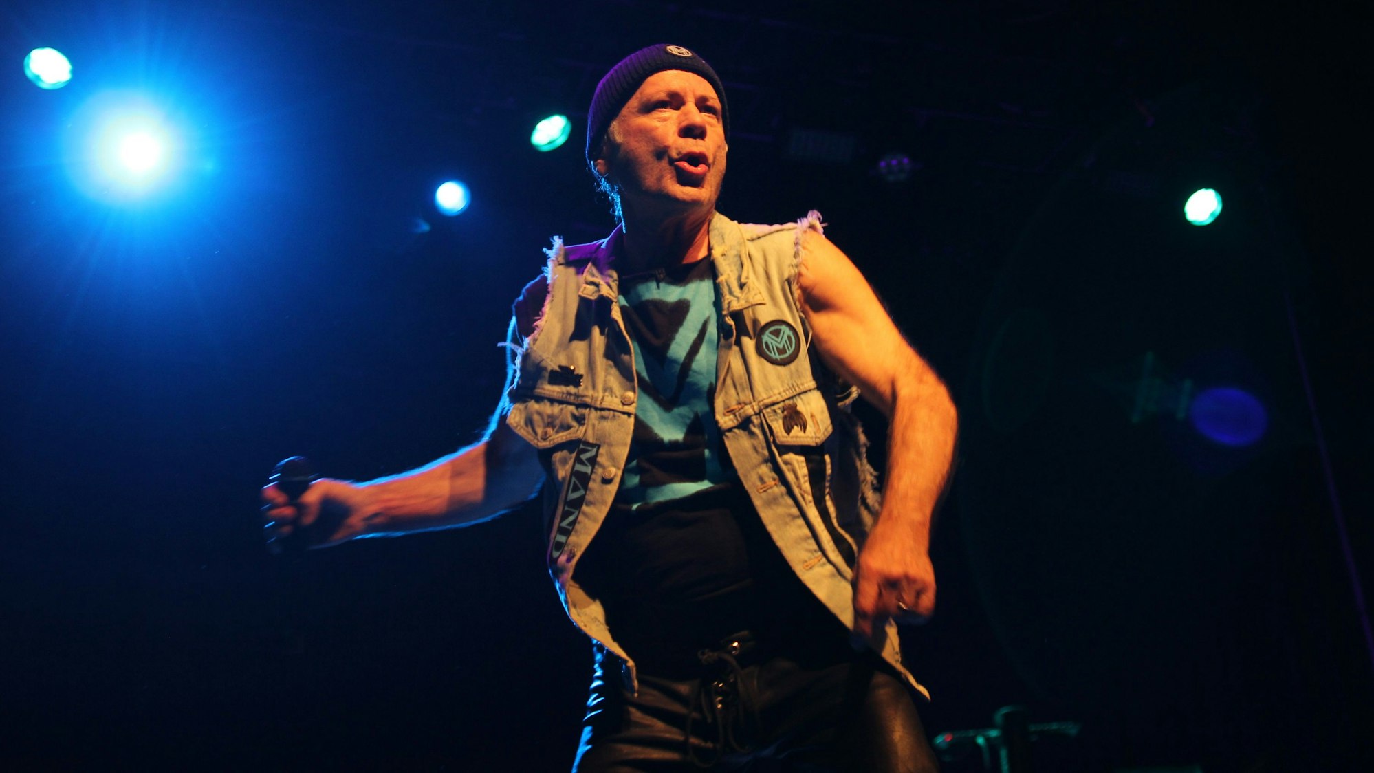 Der Sänger Bruce Dickinson steht mit einem Mikrofon in der Hand auf der Bühne. Er trägt eine Wollmütze und eine Jeansweste.