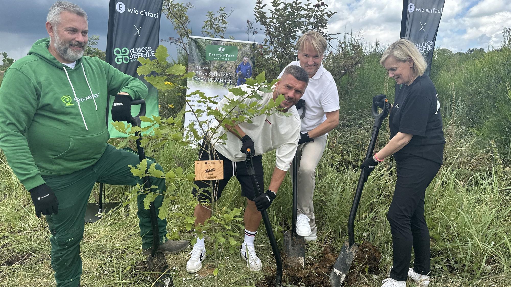 Lukas Podolski und weitere Männer und eine Frau stehen in einem Wald in Lindlar und pflanzen einen Baum.