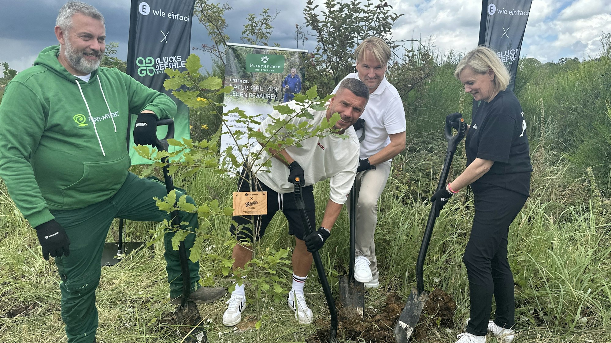 Lukas Podolski und weitere Männer und eine Frau stehen in einem Wald in Lindlar und pflanzen einen Baum.