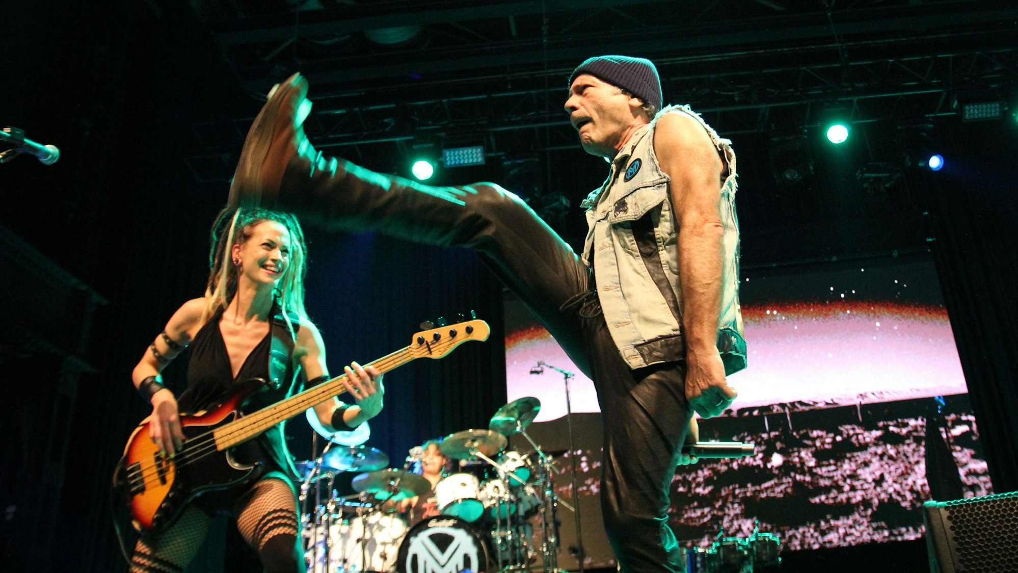 Sänger Bruce Dickinson reißt das rechte Bein in Andeutung eines Sprungs nach oben. Im Hintergrund amüsiert sich Bassistin Tanya O’Callaghan darüber.