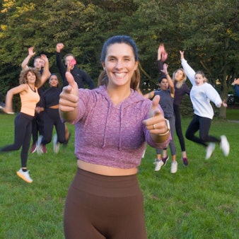 Eine junge Frau steht in einem Park und lacht in die Kamera. Hinter ihr springen mehrere junge Frauen in die Luft.
