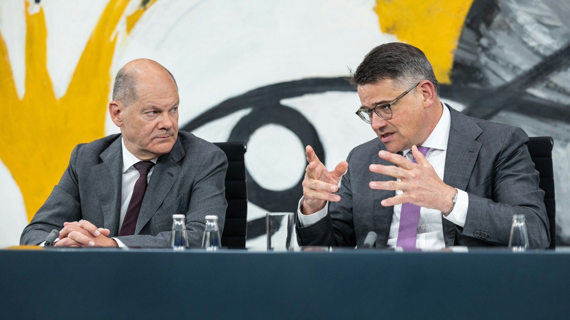 Bundeskanzler Scholz (SPD) und Boris Rhein (CDU), Ministerpräsident von Hessen, sitzen während einer PK zur Ministerpräsidentenkonferenz am 20. Juni nebeneinander. Während Rhein spricht und mit den Händen gestikuliert, sitzt Scholz mit gefalteten Händen und schaut Rhein an. Den Bildhintergrund liefert ein großflächiges Gemälde in den Farben gelb, weiß, grau und schwarz.