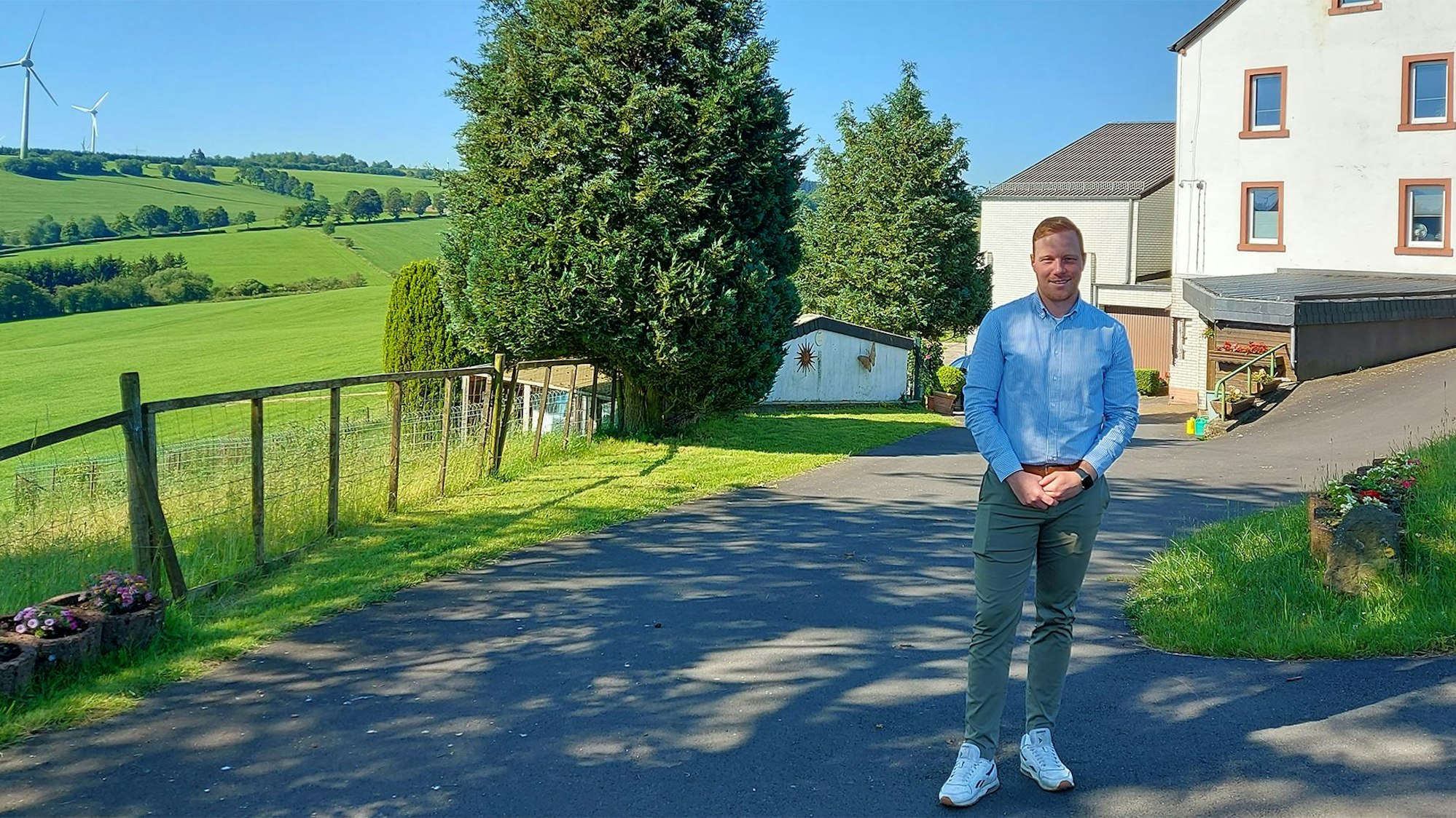Ein junger Mann steht in grauer Hose und hellblauem Hemd in der Einfahrt eines weißen Hauses. Im Hintergrund sind grüne Hügel und Windräder zu erkennen.