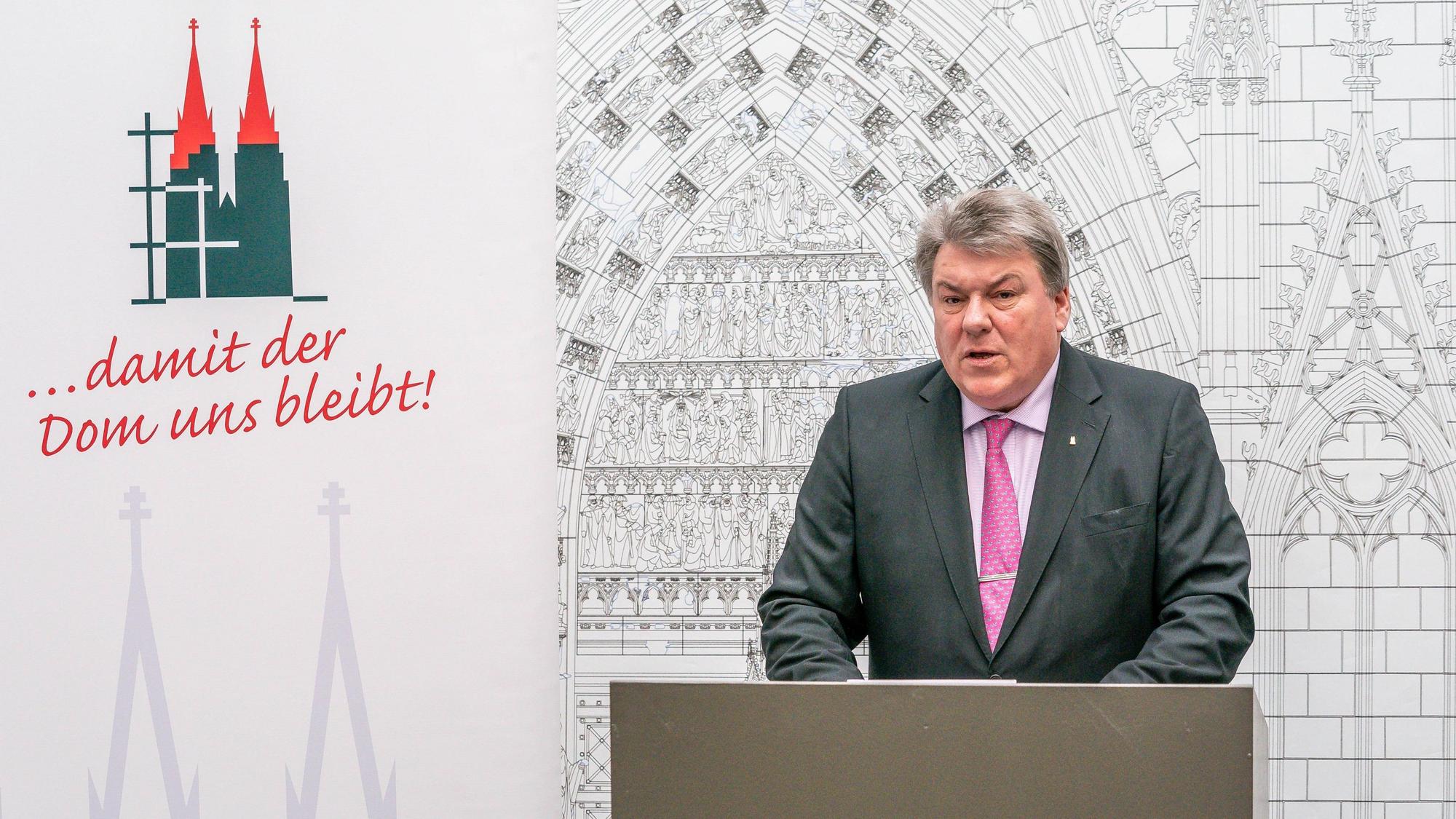 Michael Kreuzberg, Präsident des Kölner Zentral-Dombau-Vereins, steht während einer Pressekonferenz an einem Rednerpult. Er trägt grauen Anzug, roséfarbenes Hemd und rosa Krawatte. Den Hintergrund rechts füllt eine Zeichnung vom Eingangsportal des Doms aus, links ist ein rot-grünes Logo des Doms mit der Unterschrift "... damit der Dom uns bleibt!" zu sehen.