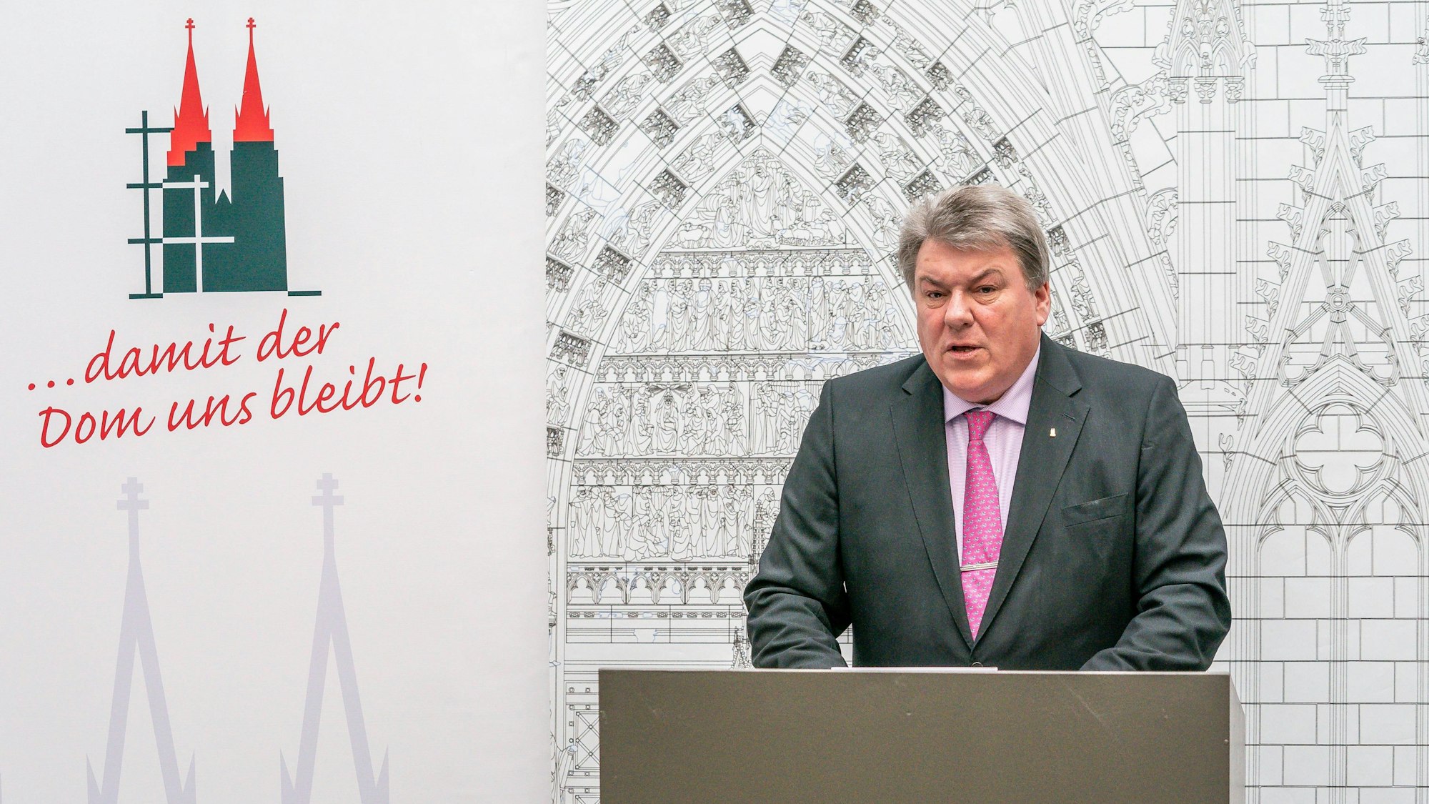 Michael Kreuzberg, Präsident des Kölner Zentral-Dombau-Vereins, steht während einer Pressekonferenz an einem Rednerpult. Er trägt grauen Anzug, roséfarbenes Hemd und rosa Krawatte. Den Hintergrund rechts füllt eine Zeichnung vom Eingangsportal des Doms aus, links ist ein rot-grünes Logo des Doms mit der Unterschrift "... damit der Dom uns bleibt!" zu sehen.