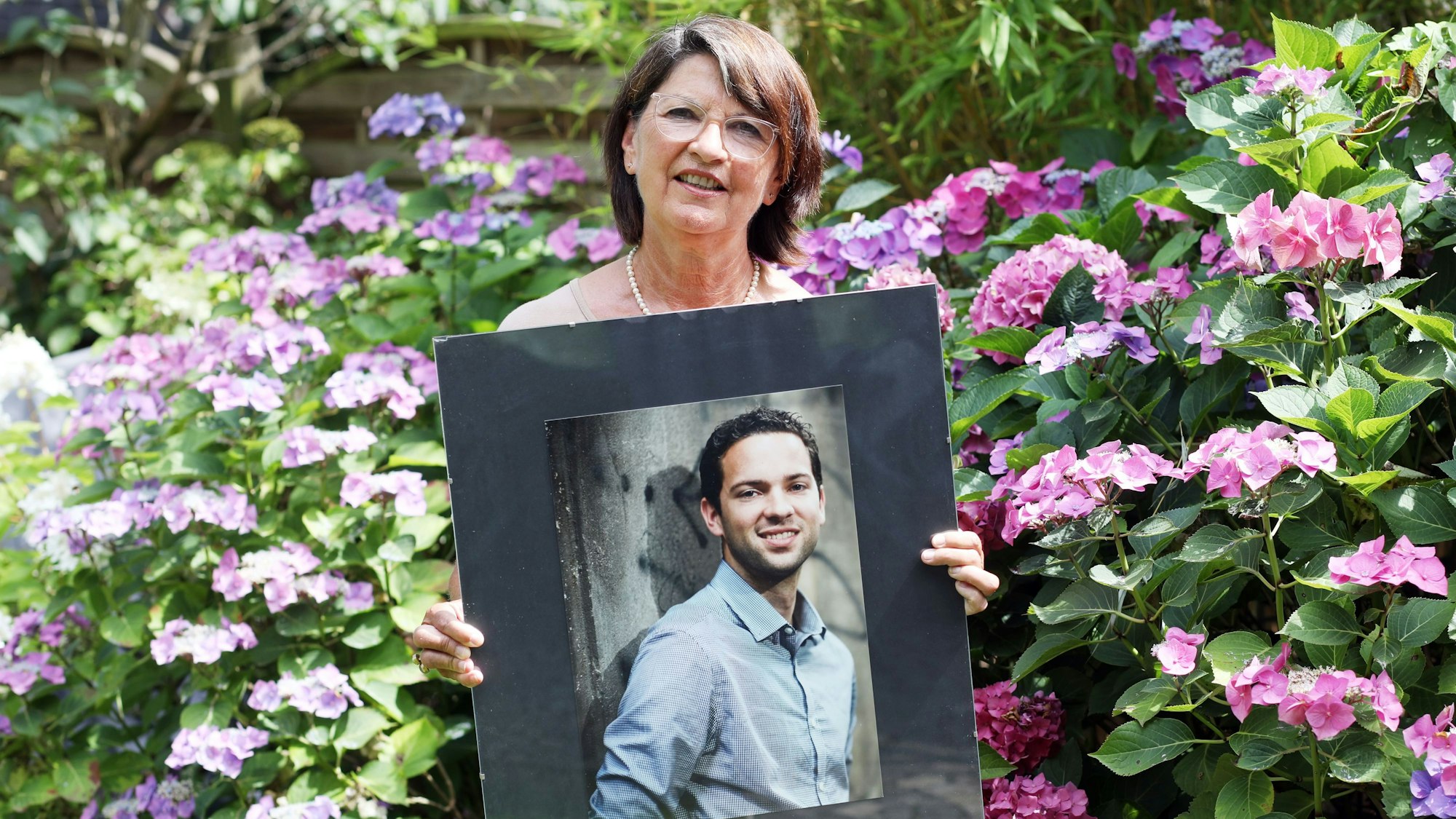 Bernadette Rüggeberg zeigt ein großes Porträt ihres Sohnes, im Hintergrund blühende Hortensien