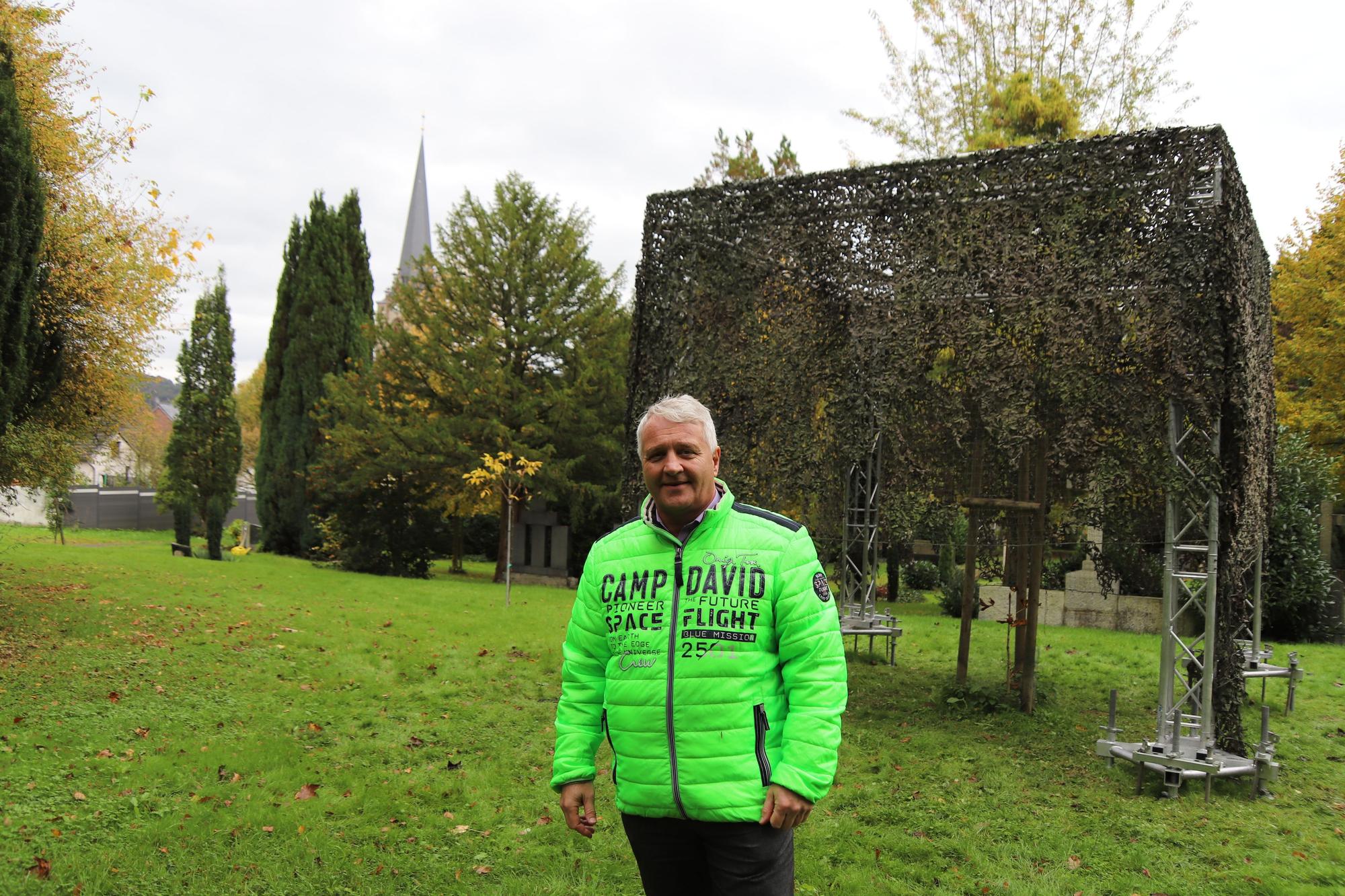 Der Bürgermeister Rainer Viehof mit leuchtend grüner Jacke steht vor einem Modell eines Memorial Cube aus einem Gerüst und darüber gehängten Tarnnetz.