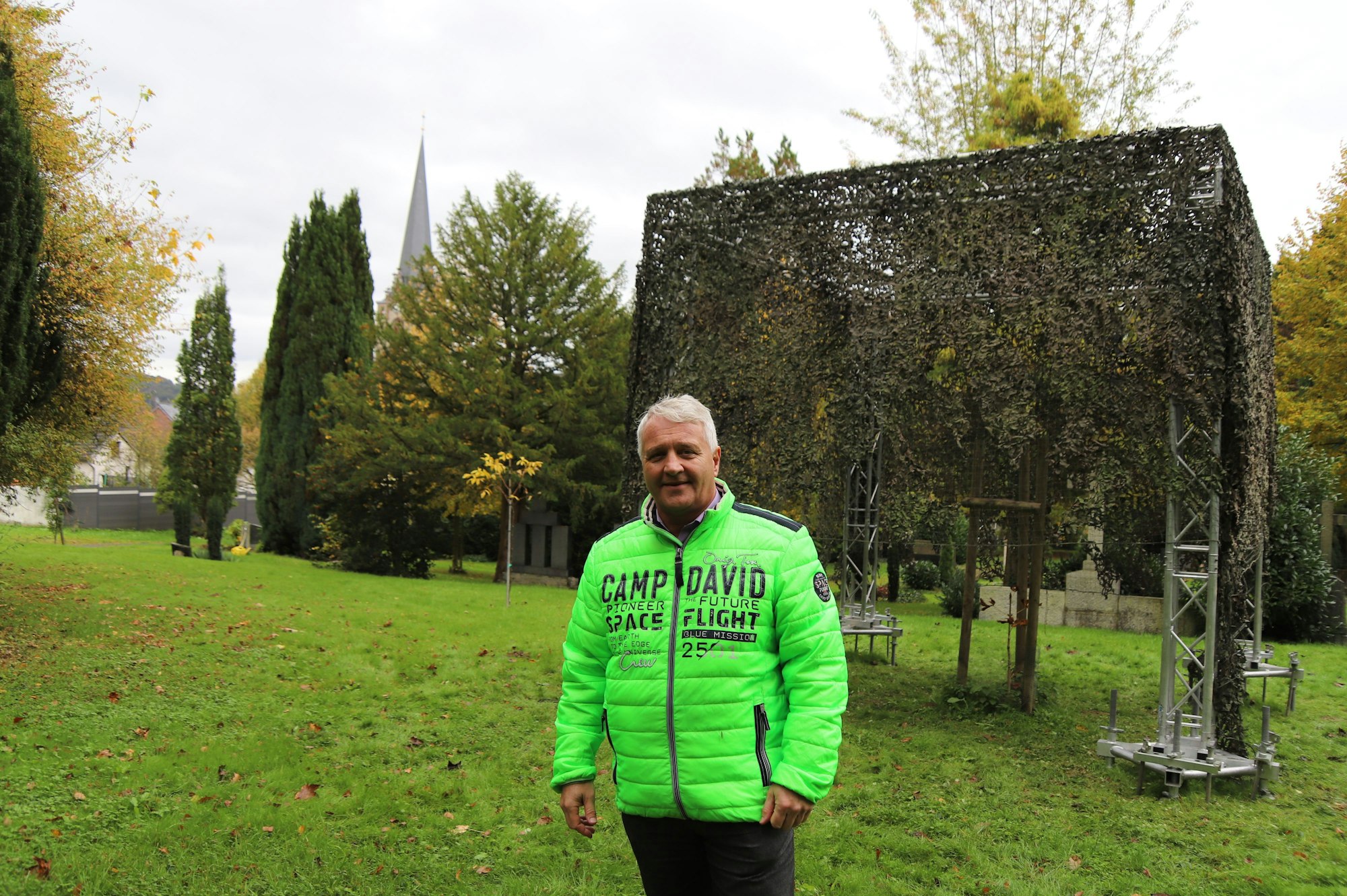 Der Bürgermeister Rainer Viehof mit leuchtend grüner Jacke steht vor einem Modell eines Memorial Cube aus einem Gerüst und darüber gehängten Tarnnetz.