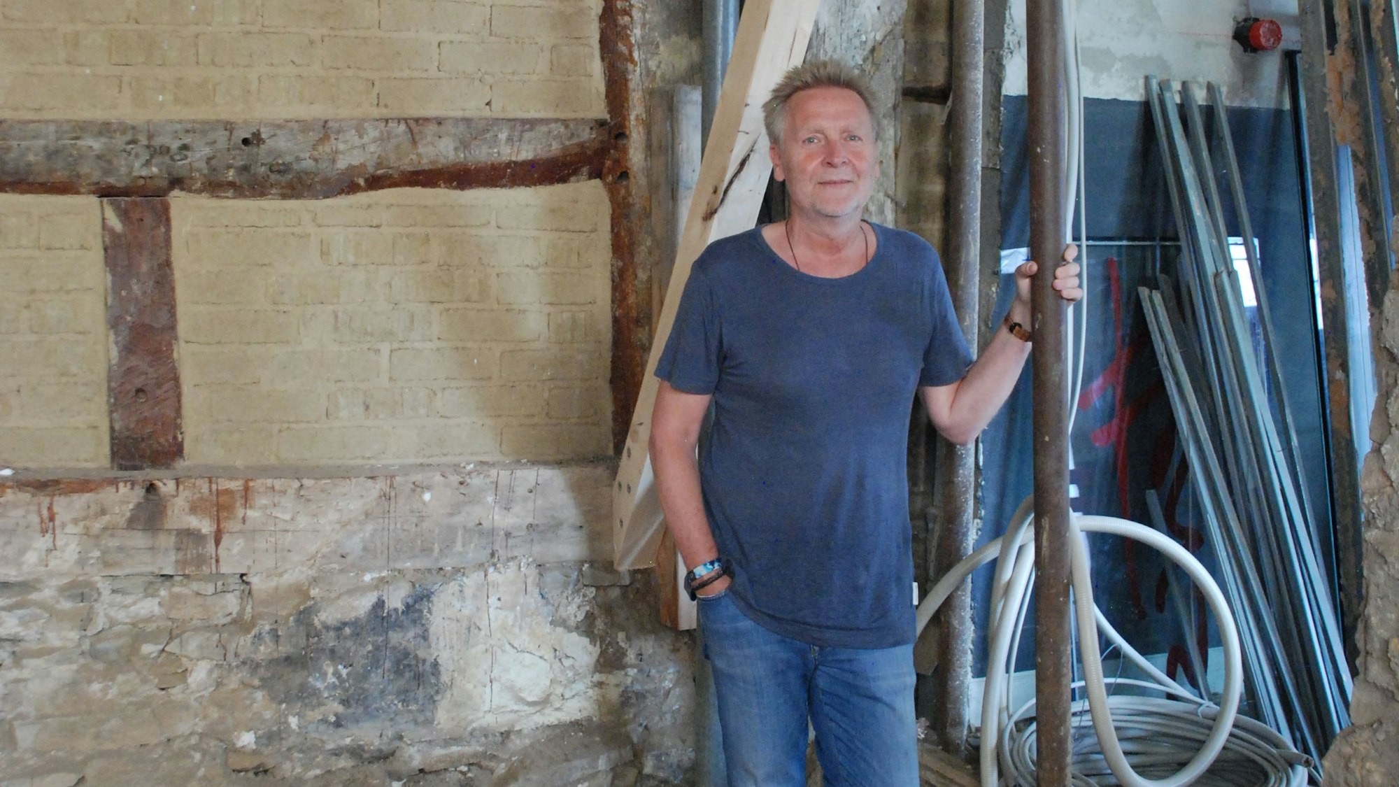 Josef Mütters steht inmitten einer Baustelle. Er hält sich an einem Rohr fest. Im Hintergrund ist eine nackte Steinwand zu sehen.