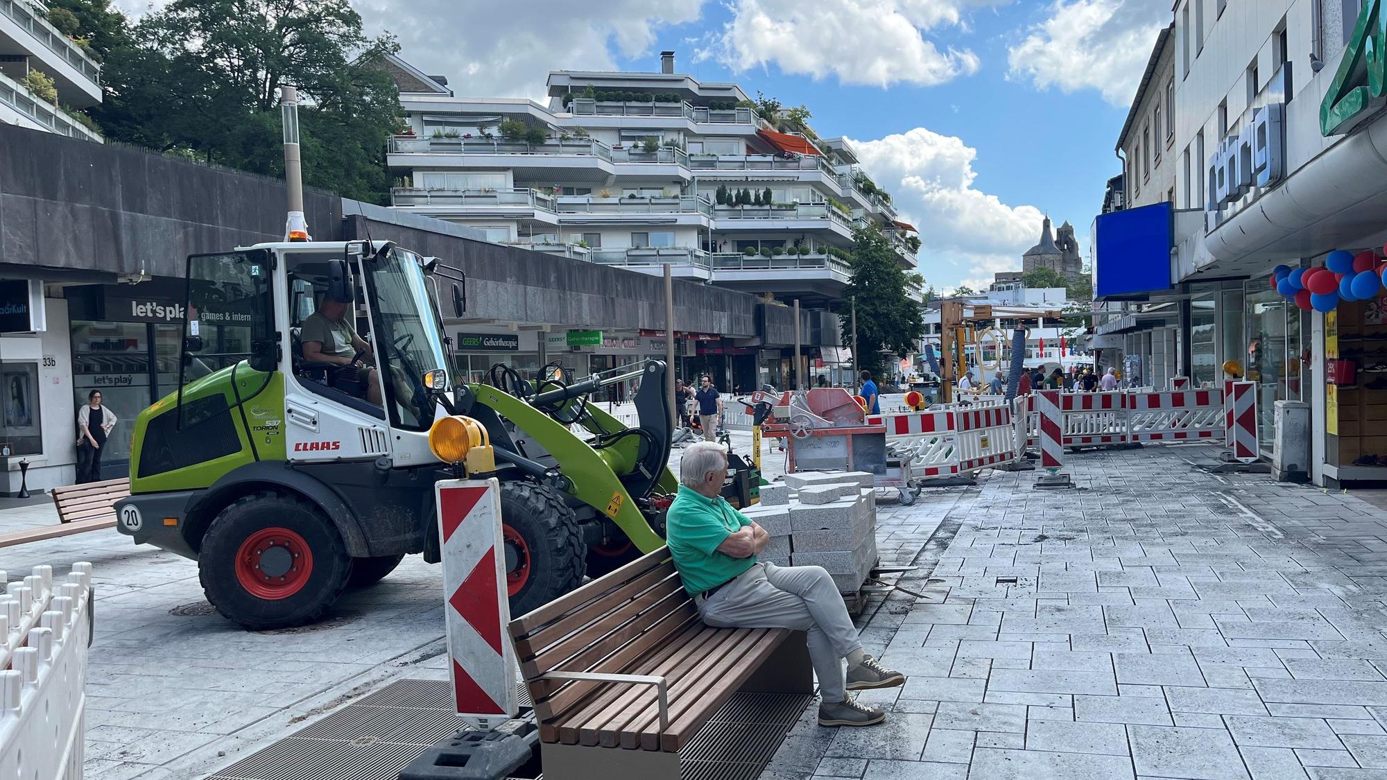 Ein Mann sitzt auf einer Bank, um ihn herum Baustellenabsperrungen, hinter ihm ein grüner Bagger.