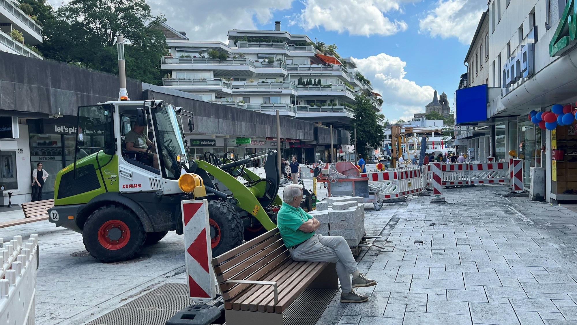 Ein Mann sitzt auf einer Bank, um ihn herum Baustellenabsperrungen, hinter ihm ein grüner Bagger.