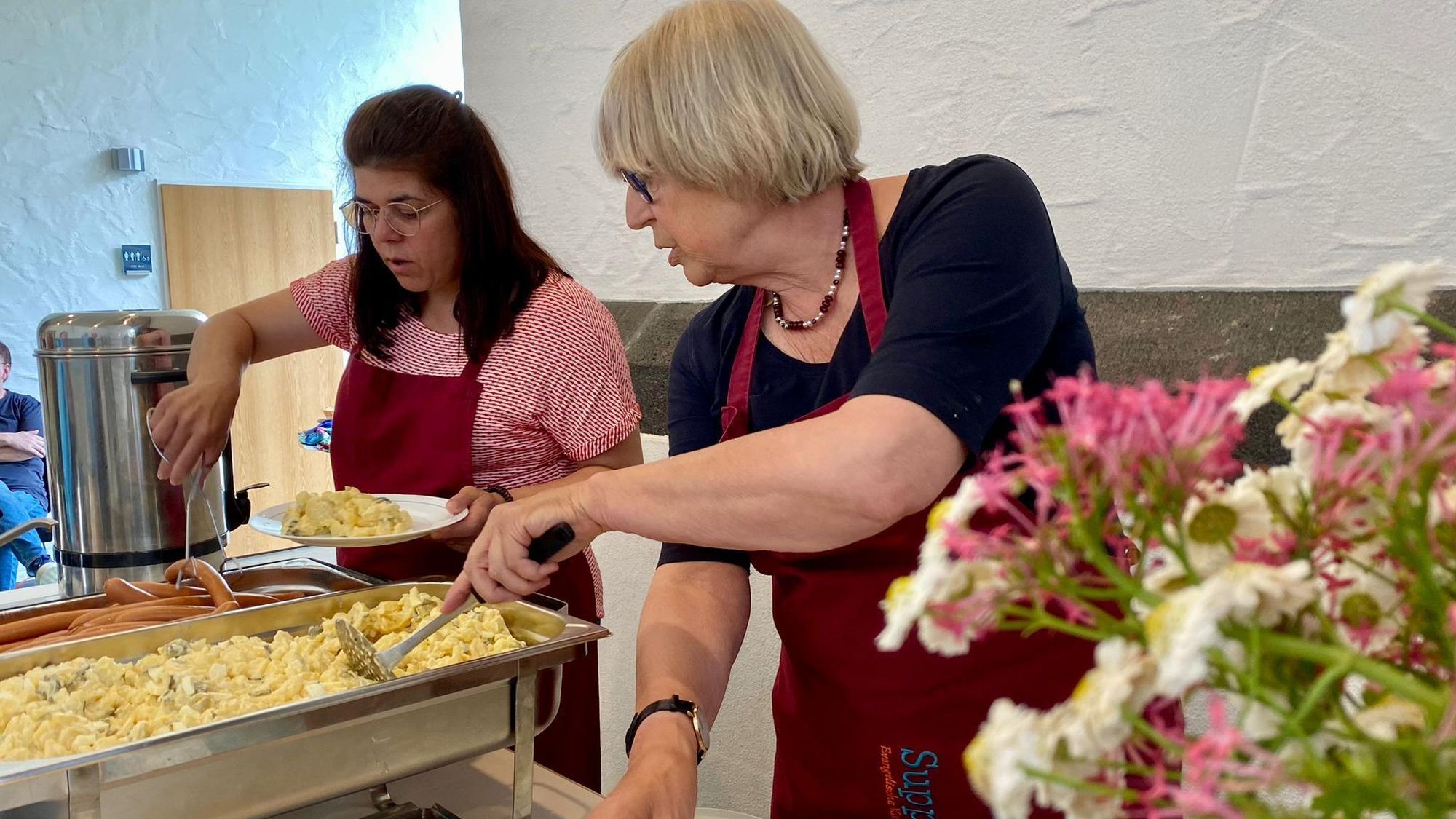 Zwei ehrenamtliche Mitarbeiterinnen verteilen Kartoffelsalat und Würstchen auf Teller.