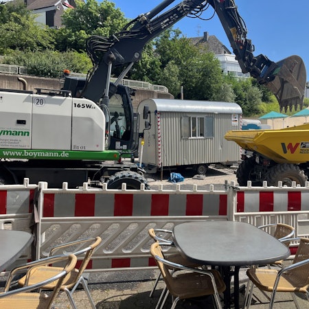 Unmittelbar vor der Absperrung stehen Tische eines Restaurants. Unmittelbar dahinter fährt ein Bagger.