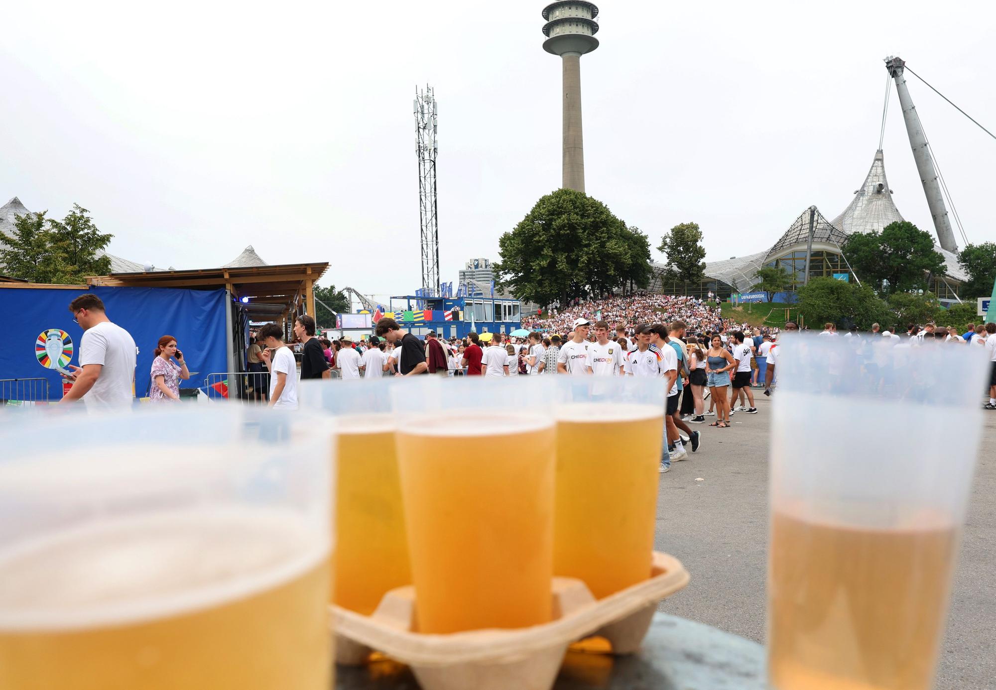 29.06.2024, Bayern, München: Fußball: EM, Deutschland - Dänemark, Public Viewing München. Fans stehen in der Fanzone im Olympiapark hinter Bechern mit Bier in einer Schlange vor einem Bierstand.