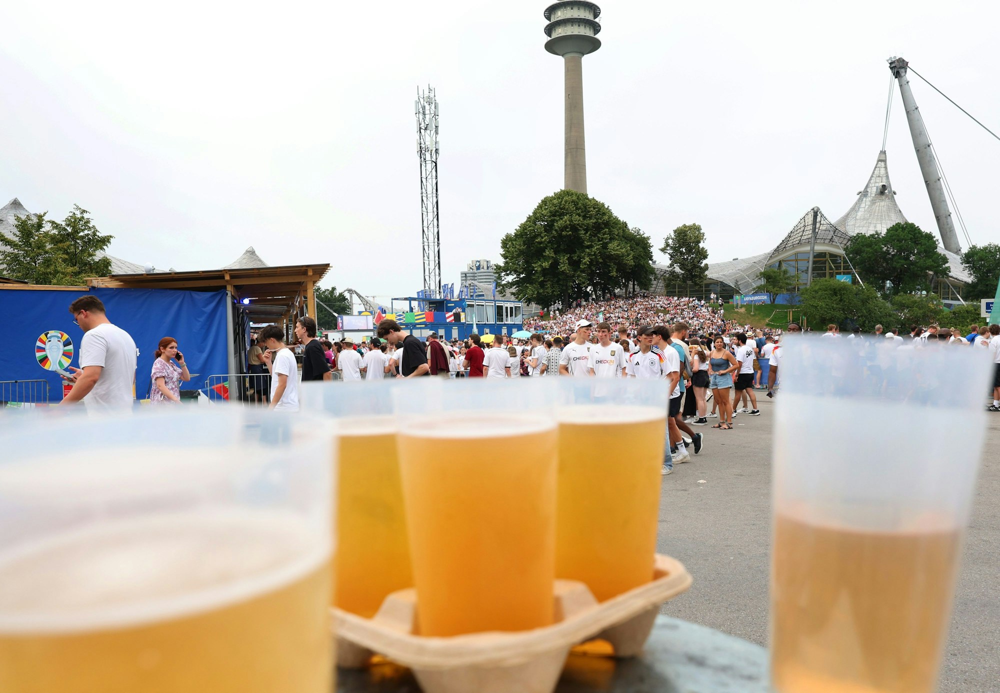 29.06.2024, Bayern, München: Fußball: EM, Deutschland - Dänemark, Public Viewing München. Fans stehen in der Fanzone im Olympiapark hinter Bechern mit Bier in einer Schlange vor einem Bierstand.