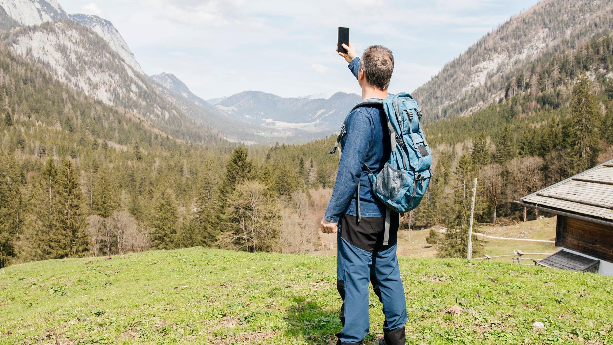 Ein Wanderer steht mit einem Smartphone auf einer kleinen Wiese in den Bergen