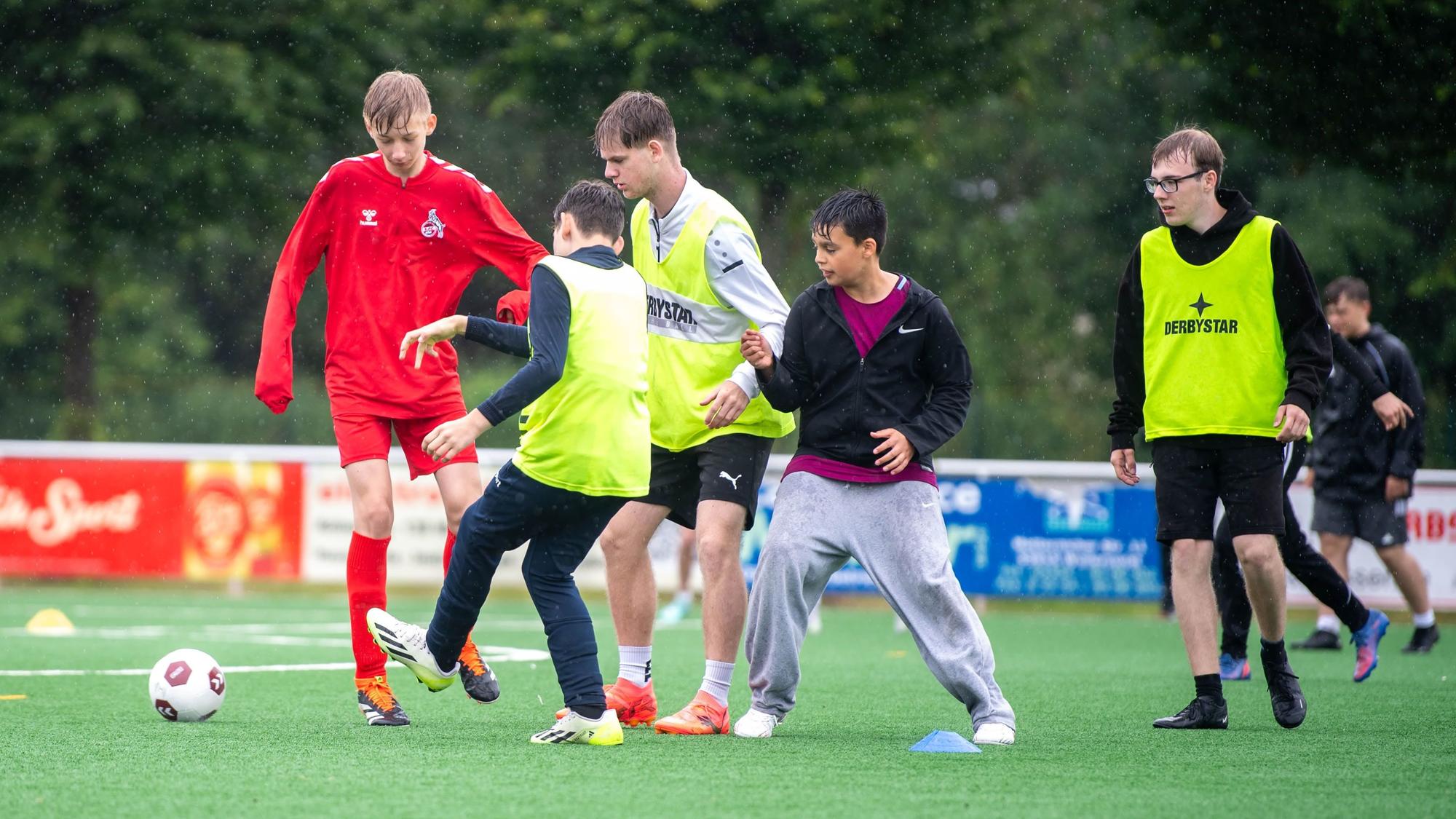 Das Bild zeigt eine Gruppe von Jugendlichen, die auf dem Kunstrasen in Weilerswist im Regen Fußball spielen.