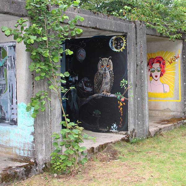 Auf den Wänden von Garagen ähnlichen Beton-Elementen sind Bilder gemalt. Ein Bild zeigt eine rothaarige Frau, ein anderes eine Eule. Um die Betonelemente herum sind Wiese und Bäume.