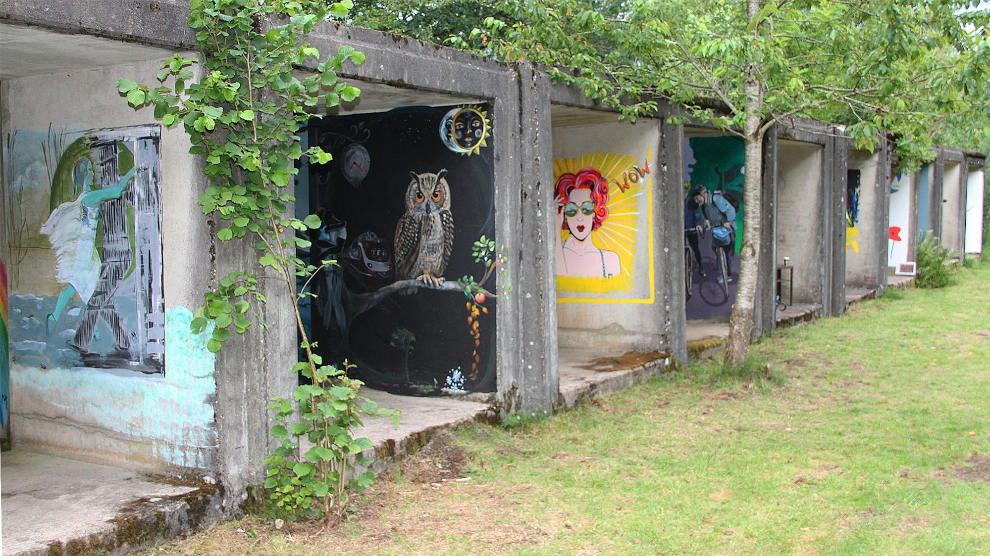 Auf den Wänden von Garagen ähnlichen Beton-Elementen sind Bilder gemalt. Ein Bild zeigt eine rothaarige Frau, ein anderes eine Eule. Um die Betonelemente herum sind Wiese und Bäume.
