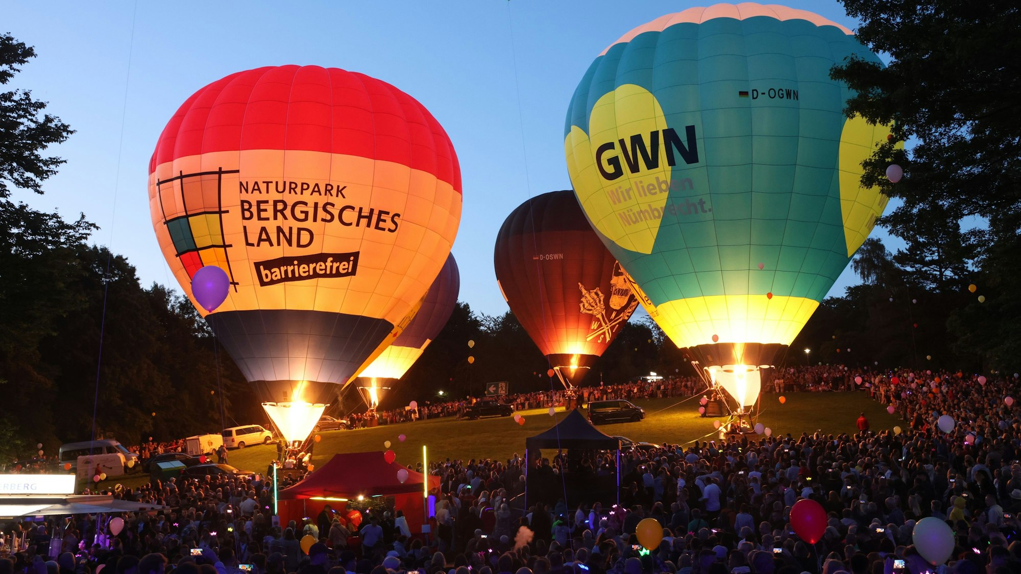 Vier Heißluftballone glühen in der Dämmerung. Tausende sehen zu.