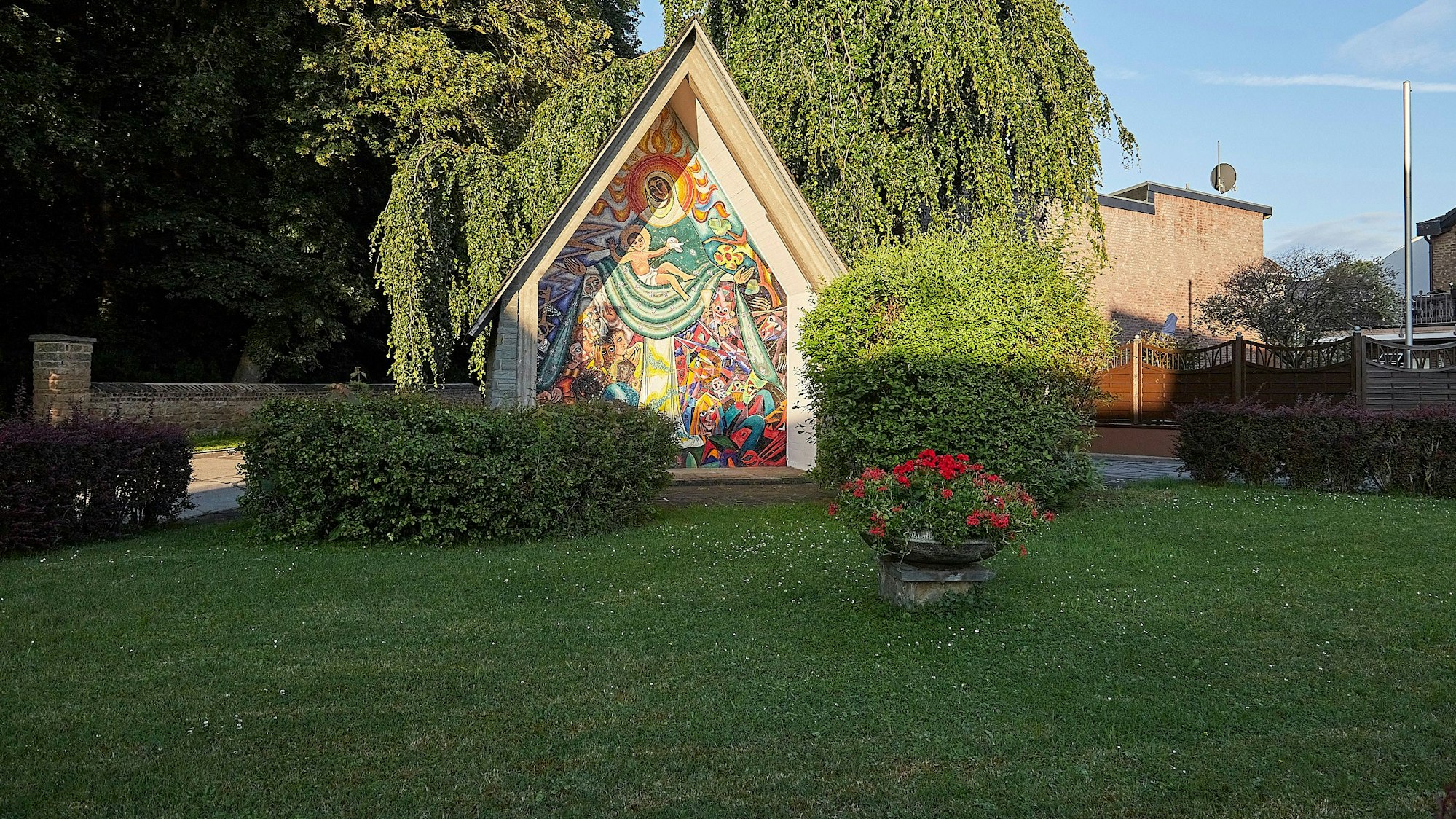 Hinter einer grünen Wiese in Vlatten ist ein Häuschen mit der Darstellung der Schutzmantelmadonna zu sehen.