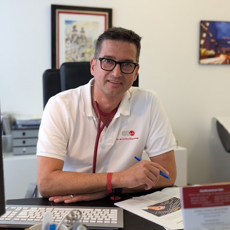 Das Foto zeigt den Arzt im weißen Poloshirt und mit Stethoskop um den Hals und Stift in der Hand an seinem Schreibtisch.
