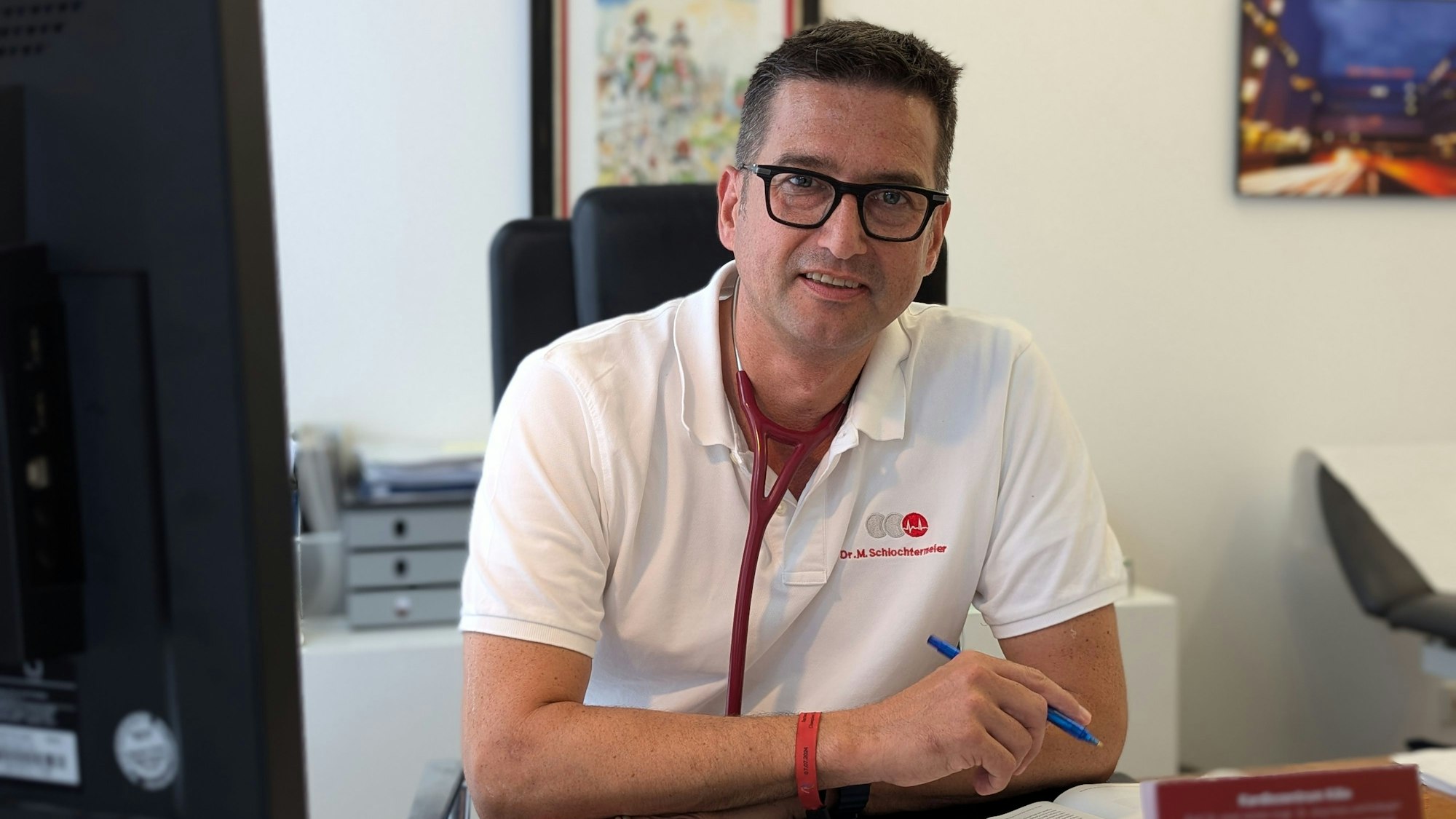 Der Arzt sitzt an seinem Schreibtisch, er trägt ein weißes Shirt und hat ein Stetoskop umgehängt.