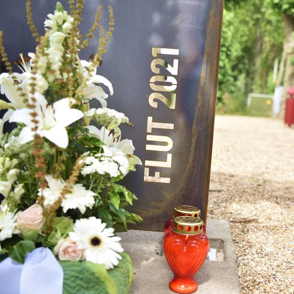 Vor einem Stahl-Schild mit der Aufschrift „Flut 2021“ stehen ein Blumengesteck und Kerzen.