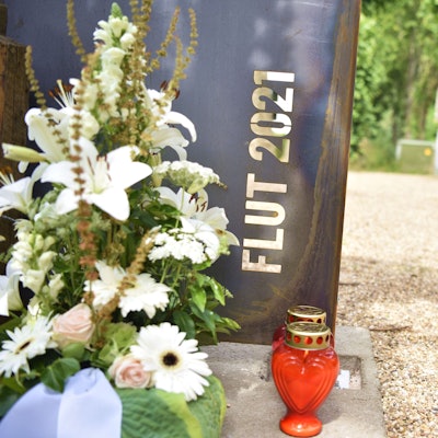 Vor einem Stahl-Schild mit der Aufschrift „Flut 2021“ stehen ein Blumengesteck und Kerzen.