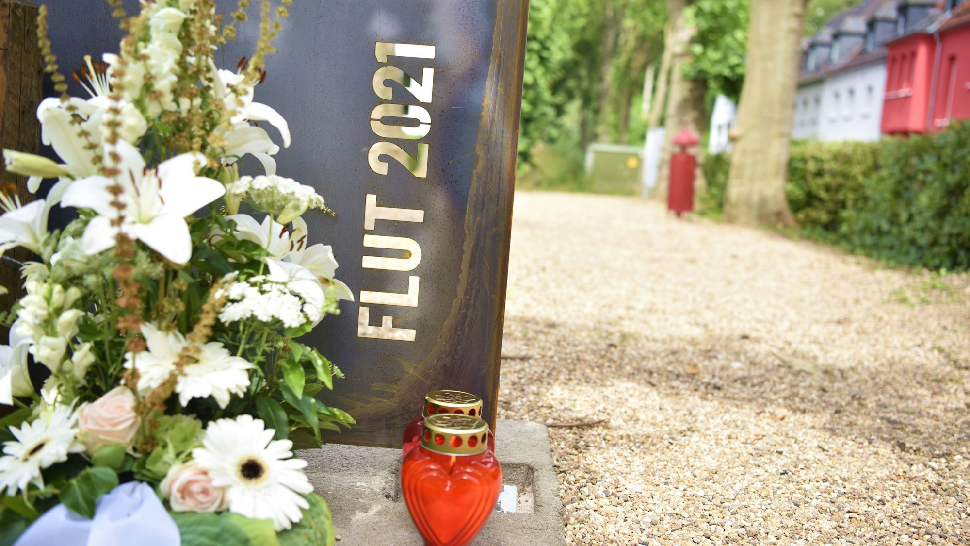 Vor einem Stahl-Schild mit der Aufschrift „Flut 2021“ stehen ein Blumengesteck und Kerzen.