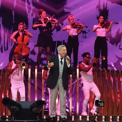 Roland Kaiser steht auf der Bühne, er trägt einen grauen Anzug mit schwarzem Jackett, im Hintergrund spielen Bläser und Streicher