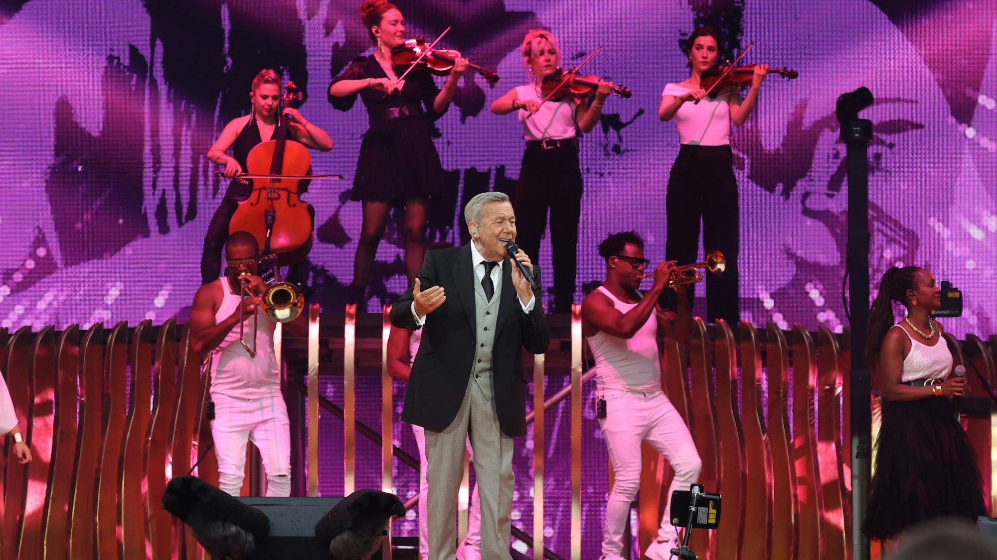 Roland Kaiser steht auf der Bühne, er trägt einen grauen Anzug mit schwarzem Jackett, im Hintergrund spielen Bläser und Streicher