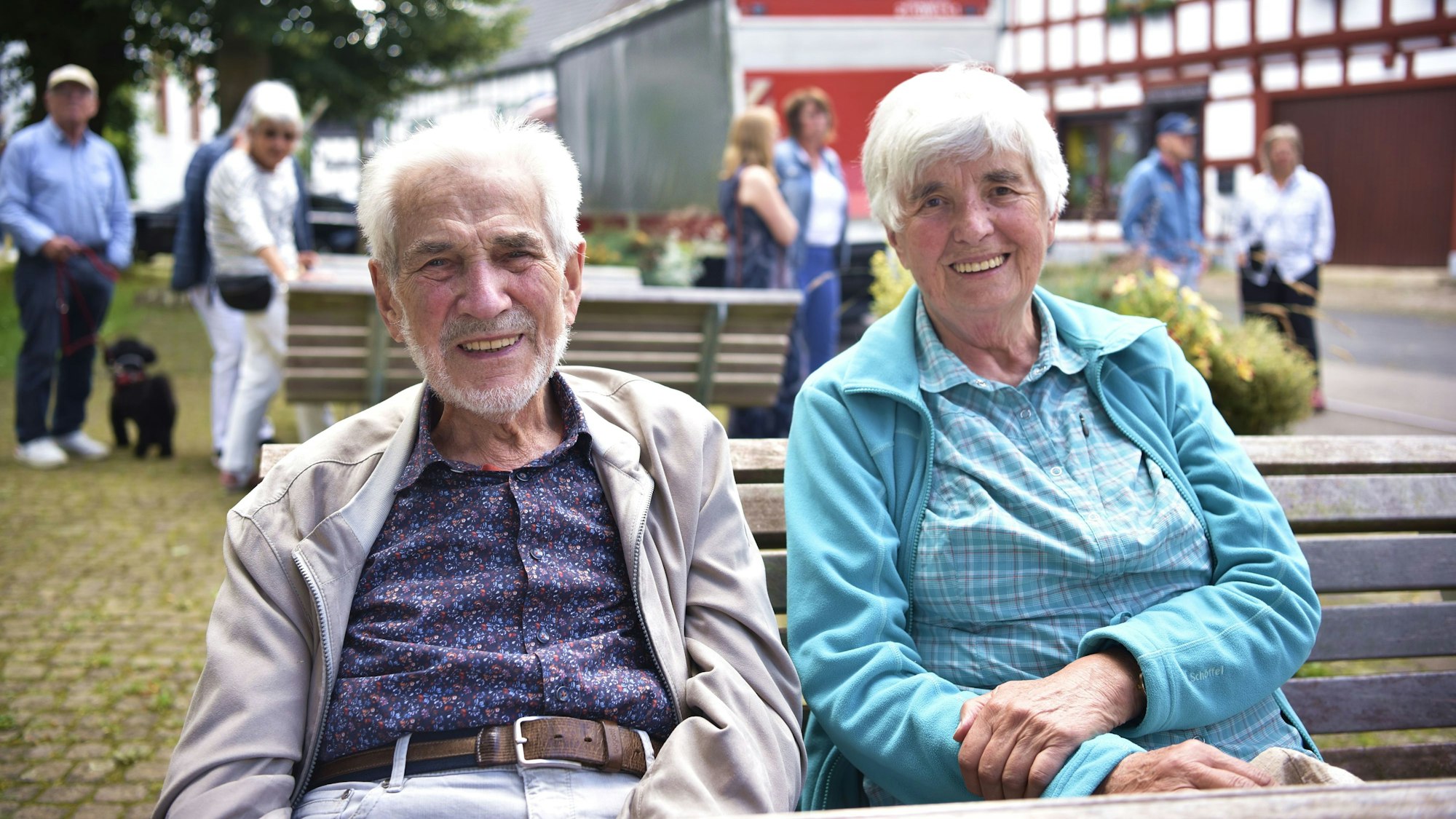 Ein Mann und eine Frau sitzen nebeneinander auf einer Bank. Im Hintergrund stehen Leute.