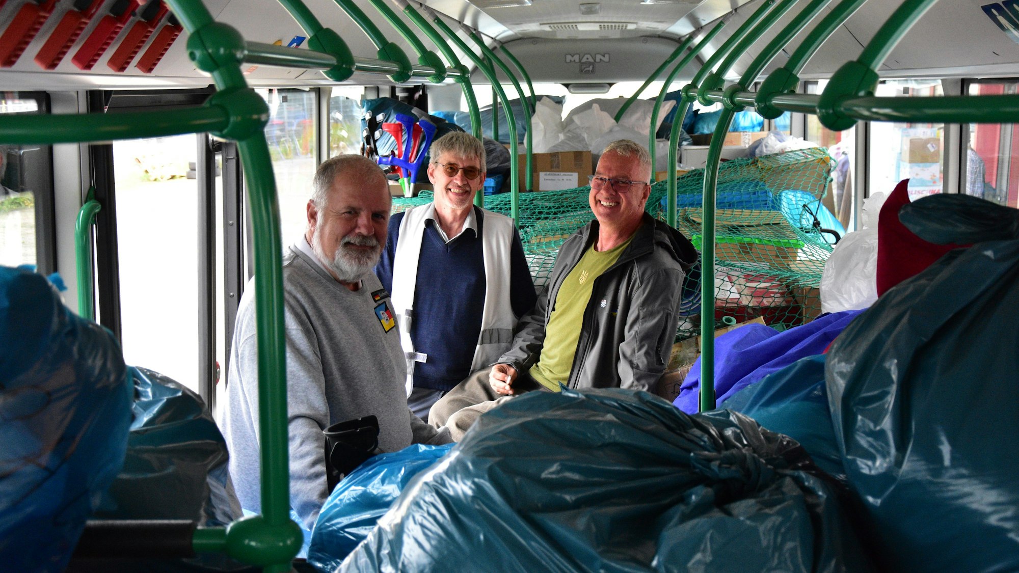 Drei Männer in einem Linienbus, in dem Plastiksäcke und Kartons gestapelt sind.