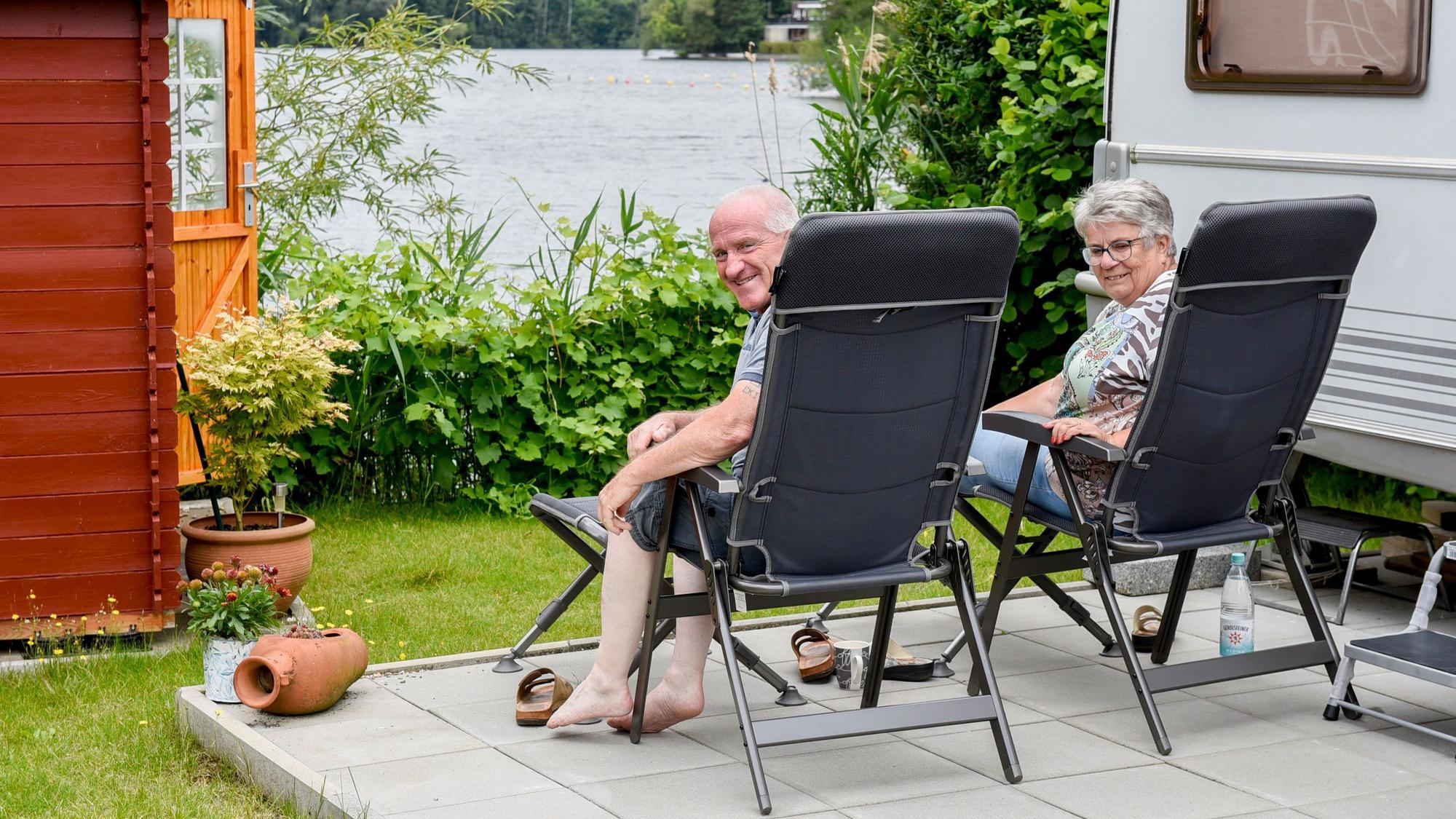 Ein Ehepaar sitzt auf Gartenstühlen an einem See neben einem Wohnwagen.