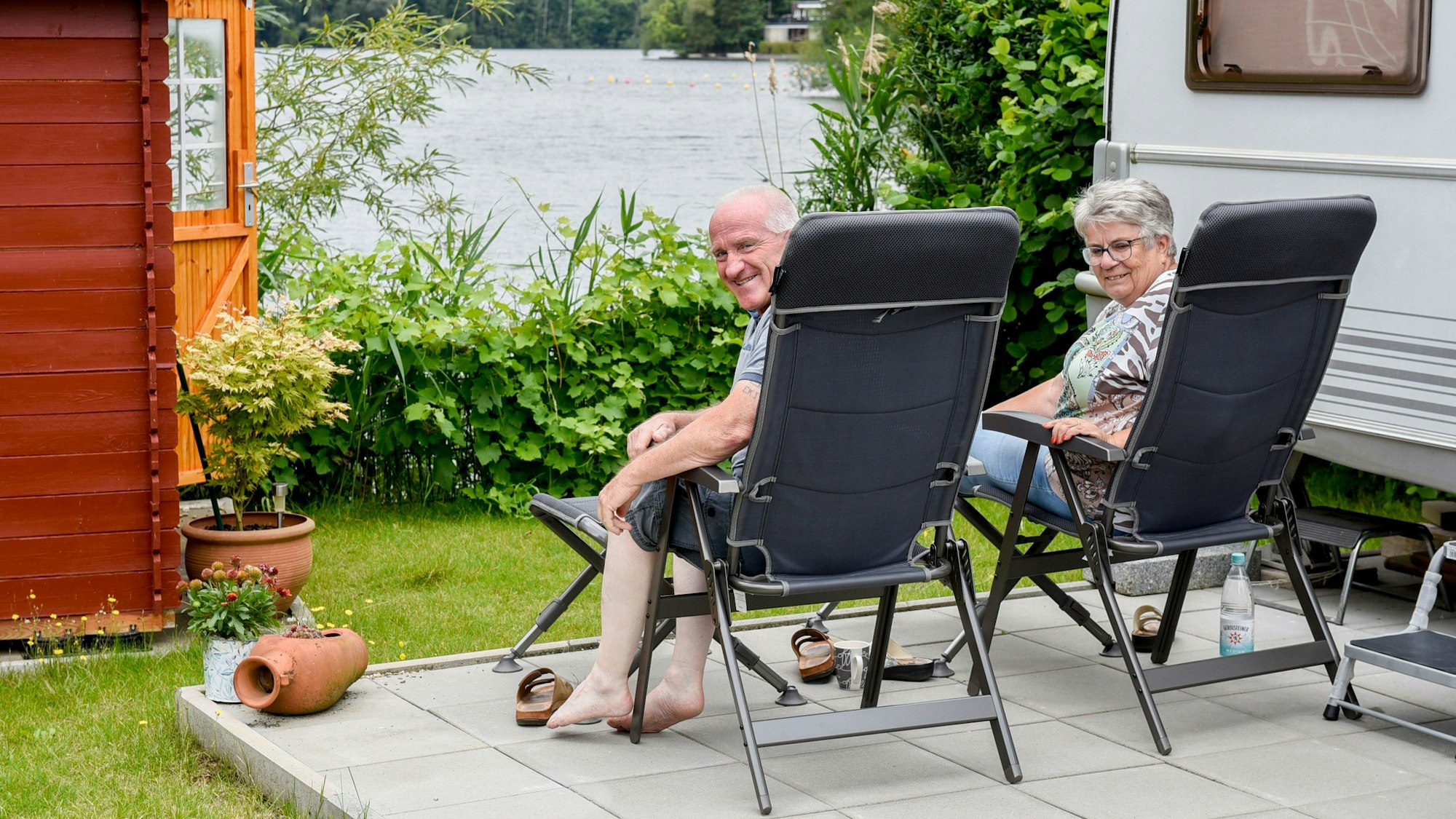 Ein Ehepaar sitzt auf Gartenstühlen an einem See neben einem Wohnwagen.