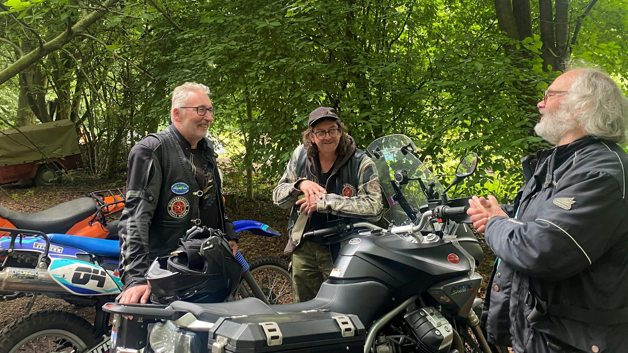 Drei Männer stehen an Motorrädern und unterhalten sich.