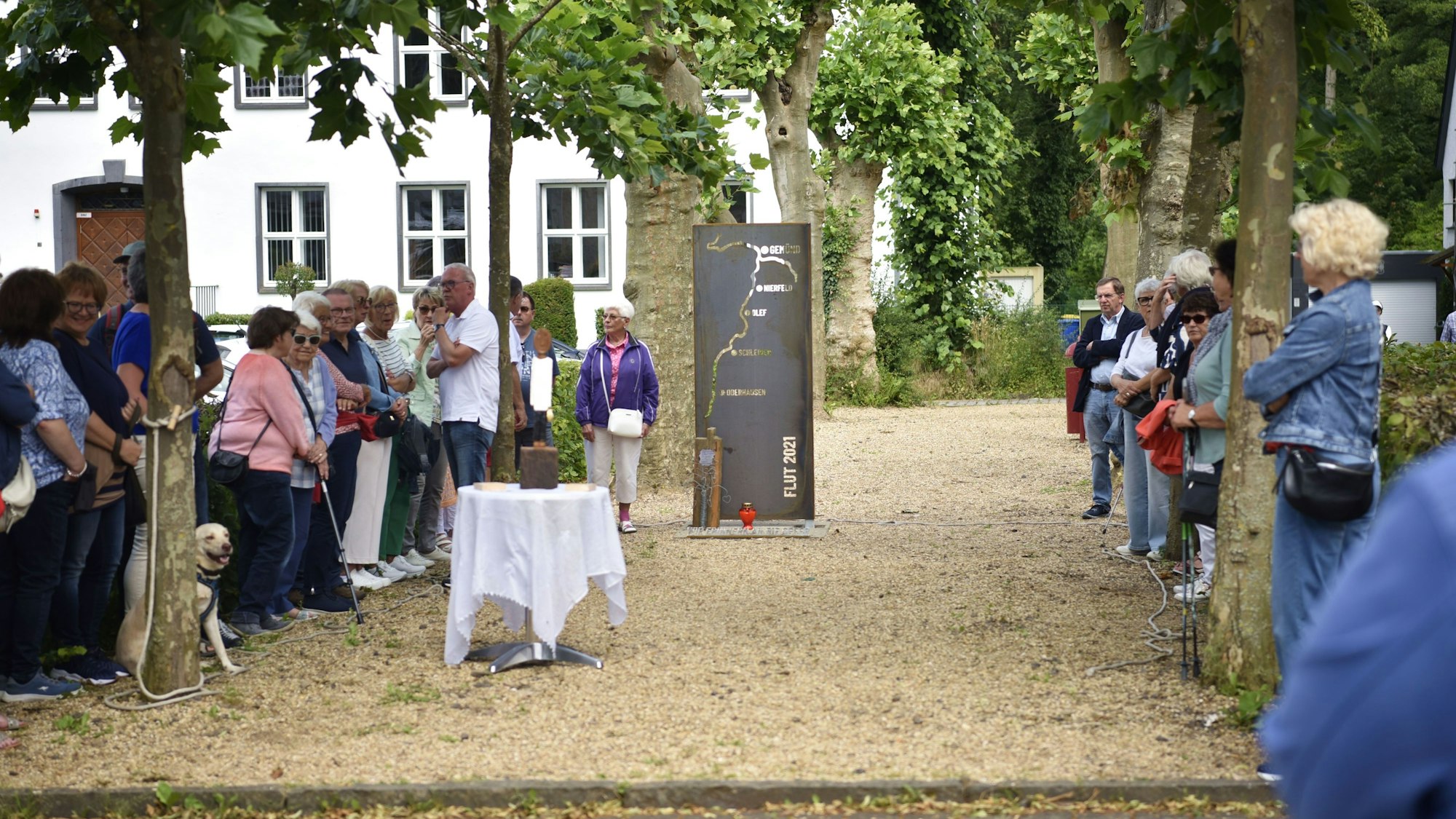 Mehrere Menschen stehen links und rechts am Rand einer Allee, in deren Mitte eine Stahl-Stele zur Flut 2021 aufgestellt ist.
