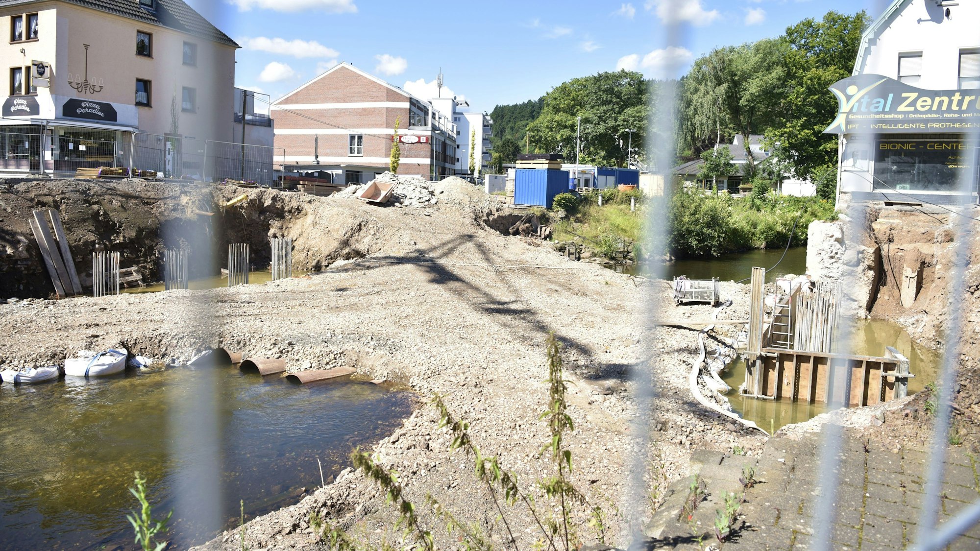 Durch einen Baustellenzaun hindurch sieht man eine Baustelle auf einem Fluss. Links und rechts sind Häuser.