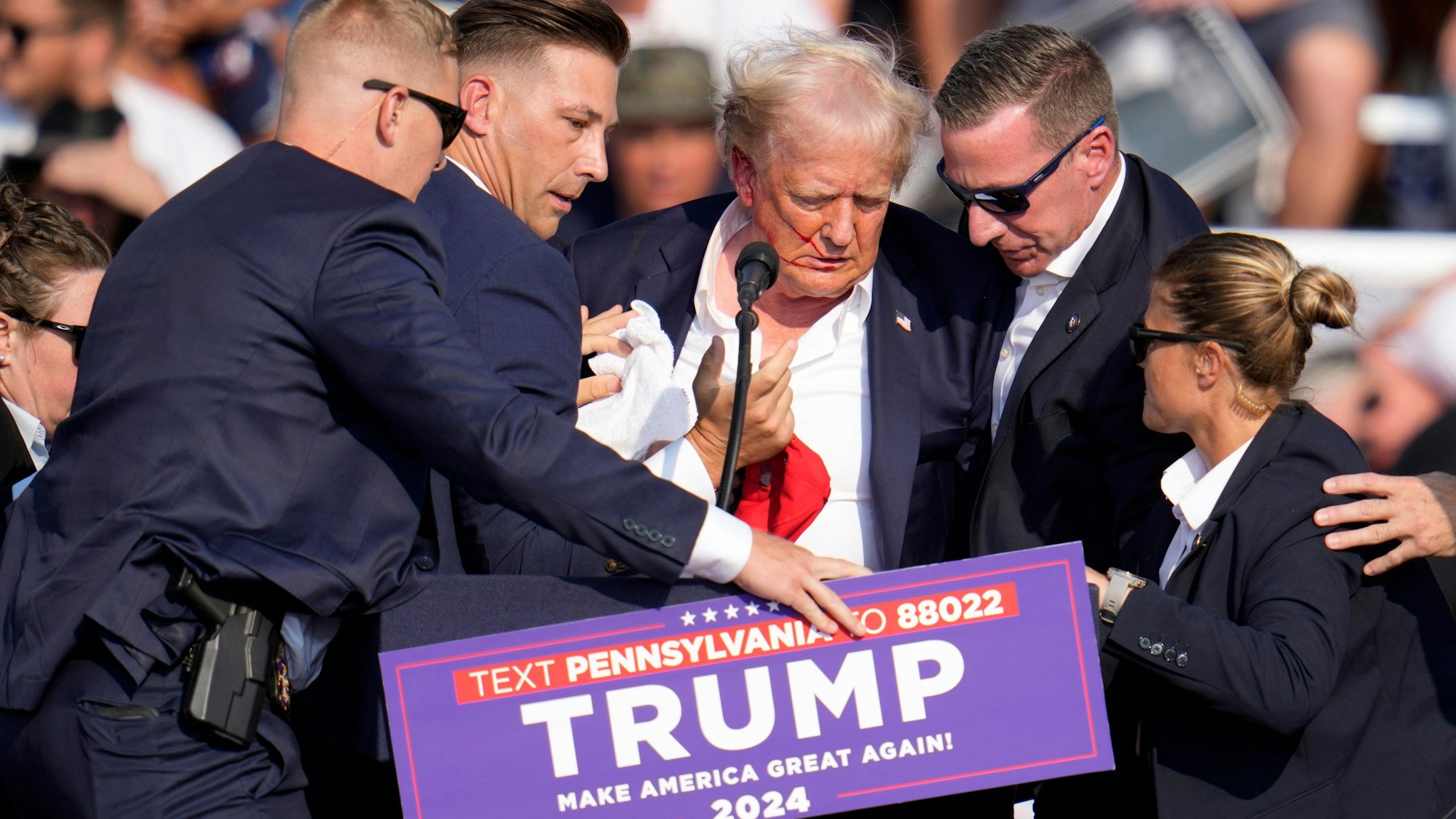 Sicherheitsbeamte schirmen den amerikanischen Ex-Präsidenten Donald Trump ab. er hat Blut im Gesicht, seine Haare sind durcheinander.